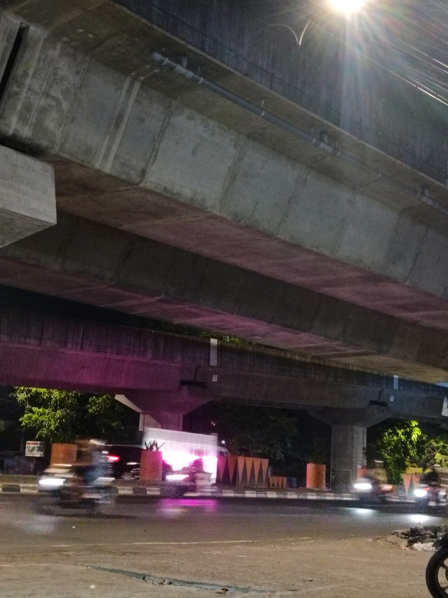 The image shows a busy street scene at night under an elevated structure, likely a highway overpass or bridge, in Makassar, Indonesia. The concrete structure above is illuminated by streetlights and a bright light source with sunburst effects, casting shadows and highlights. The road surface appears wet, reflecting the lights from vehicles and the surroundings.
Several motorcycles are visible, their lights blurred due to motion, indicating they are in motion. On the sidewalk area, there are decorative planters with geometric patterns and cylindrical orange planters. Trees with lush green foliage are visible behind the planters and the elevated structure. A white van or truck with illuminated rear lights is also present on the road. The scene is characteristic of an urban environment at night, with traffic and infrastructure elements contributing to the overall atmosphere.