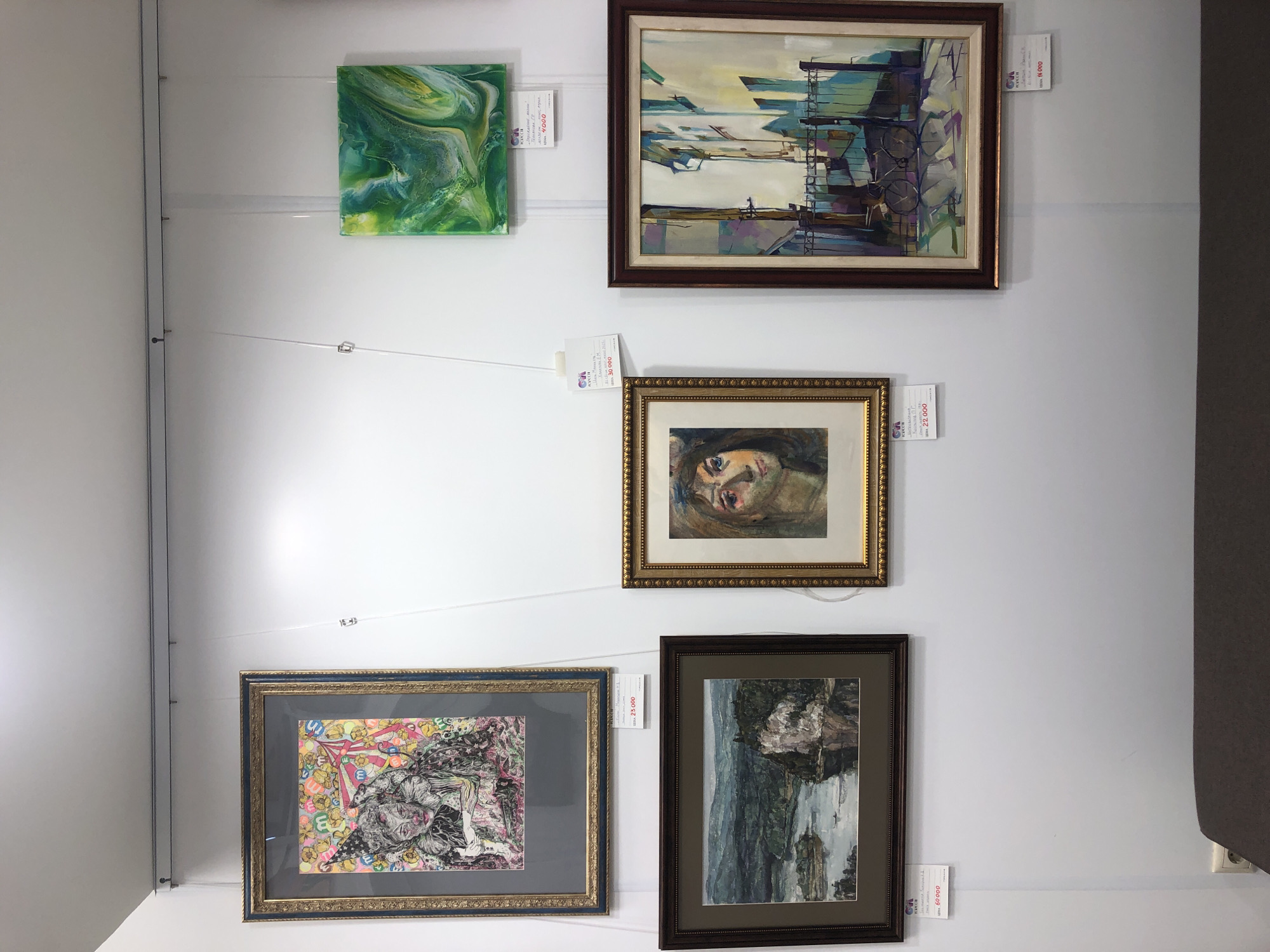 This media file displays a collection of framed artwork mounted on a white wall, likely in an art gallery or exhibition space. The artworks are displayed at varying heights, suspended by thin wires.
The subjects of the artwork include an abstract green and white fluid art piece, a landscape painting with abstract elements of buildings and structures, a portrait of a person with expressive eyes, a detailed black and white drawing overlaid with colorful elements, and a dark landscape painting of a waterfall or river with rocky cliffs.
There are several price tags visible, indicating these are items for sale. One tag, associated with the abstract green artwork, reads "4000" with some unreadable text. Another tag under the portrait painting shows "30000" and "22000". A tag below the detailed drawing indicates a price of "25000", and the landscape painting on the far right has a tag with "60000". The location context of Izhevsk, Russia, suggests this exhibition is local to that area. The lighting appears to be artificial, creating an indoor ambiance. No specific time of day or weather conditions can be inferred. The overall impression is one of an art exhibition showcasing diverse styles and subject matter.