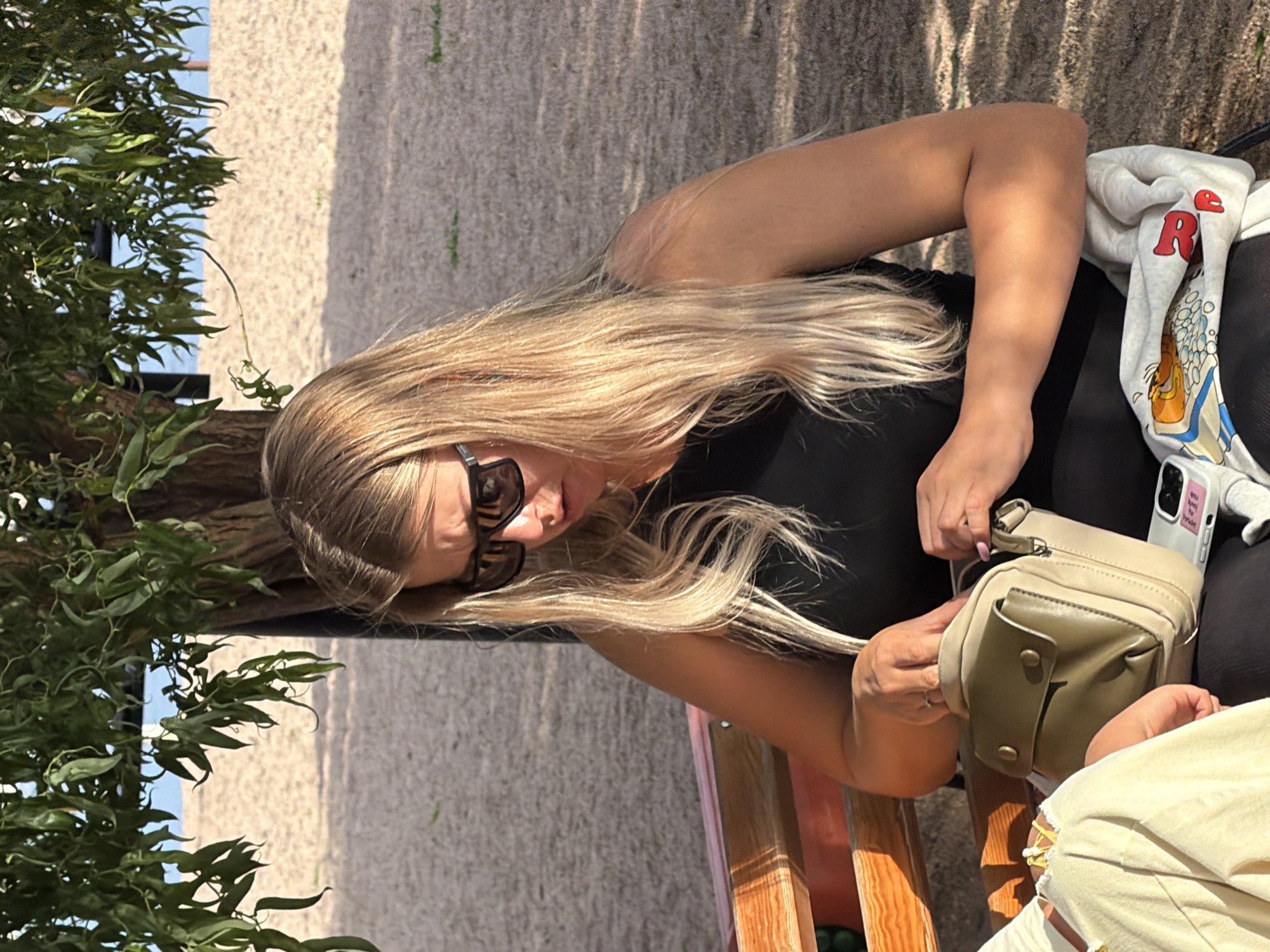 A woman with long blonde hair and sunglasses is sitting outdoors, partially obscured by a leafy tree. She is wearing a black top and is holding a light-colored purse with green accents. Her right hand is resting on the purse, and her left arm is bent at the elbow, resting on her leg. The background appears to be a gravelly or sandy surface, suggesting an outdoor setting like a park or patio. The lighting indicates it is daytime, possibly sunny, as evidenced by the shadows on the ground and the bright highlights on the woman's hair and arm. A light-colored hooded sweatshirt with red lettering is visible to her right, along with a white smartphone. The overall impression is one of a casual, relaxed moment outdoors.