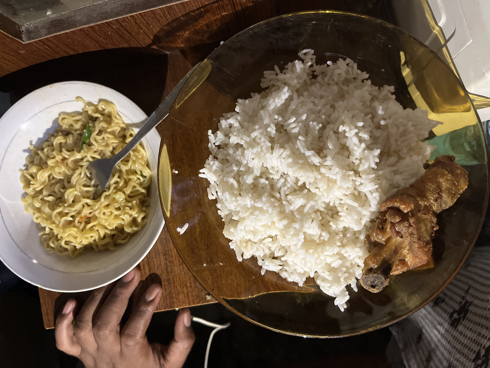 The image shows a meal laid out on a wooden surface. In the foreground, a clear, amber-colored plate holds a mound of white rice and a piece of fried chicken. Behind this, a white bowl contains a serving of instant noodles. A silver fork rests on top of the noodles, with its tines inserted into the dish. A person's hand with dark skin is visible on the left side of the frame, resting on the wooden surface near the bowl of noodles. The lighting suggests an indoor setting, possibly a dining area. The image captures a moment of a meal, likely in a home or casual eatery environment. The location in Kruengmate, Indonesia, is inferred from the context provided.