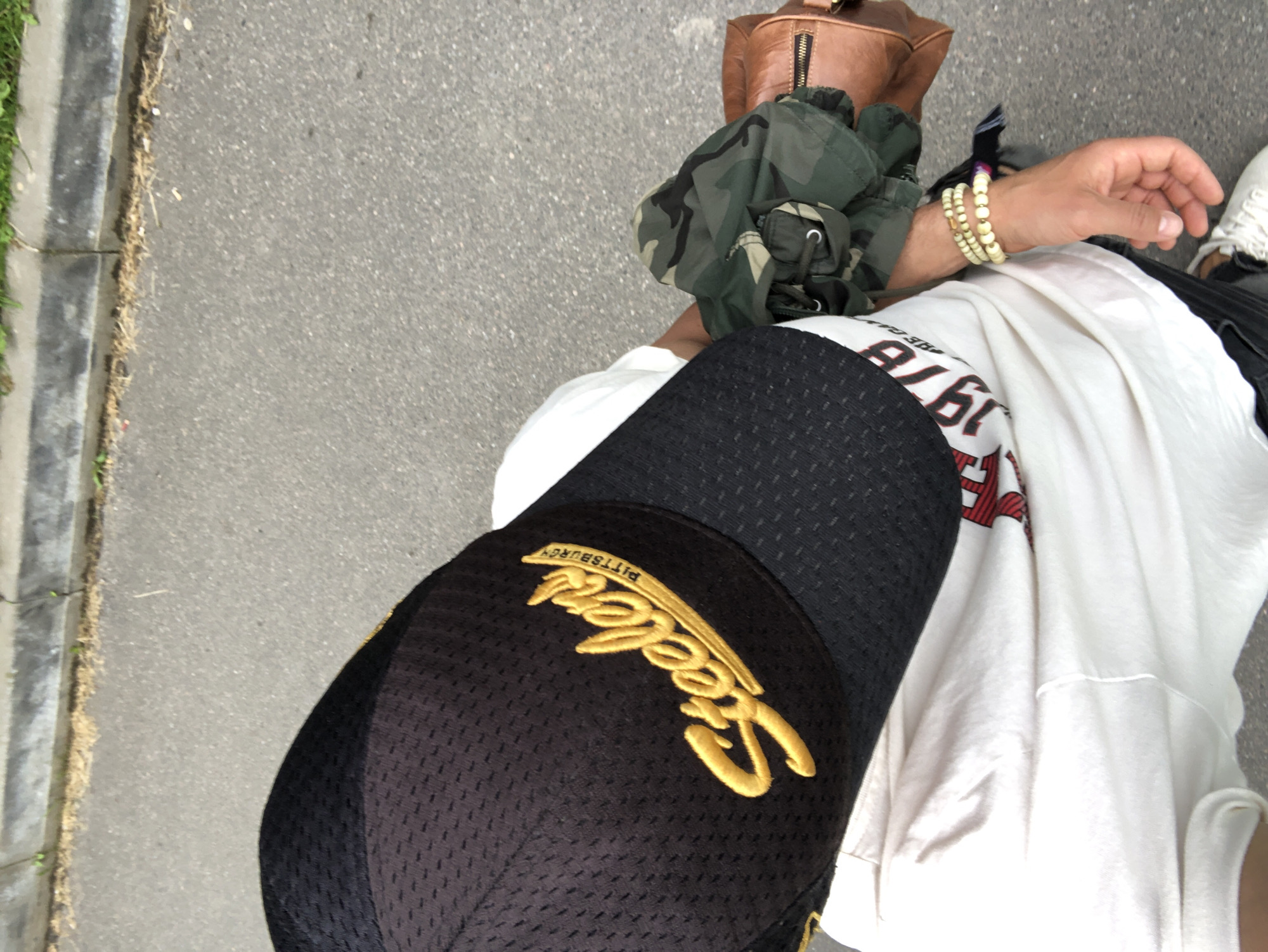 This is a medium shot, top-down perspective of a person's legs and torso, viewed from above as they stand on asphalt pavement. The person is wearing a black baseball cap with yellow embroidery, a white t-shirt with red and white graphics, a camouflage jacket, and a brown leather bag. The scene is set outdoors, with a curb and grass visible on the left side of the frame. The overall lighting suggests daytime.
The baseball cap has "PITTSBURGH" embroidered in yellow above a stylized yellow "Steelers" logo. The white t-shirt has a graphic that appears to include the numbers "1976" in red. The camouflage jacket is draped over the person's arm, and a brown leather bag with a zipper is visible near the shoulder area. The person's right wrist is adorned with two beaded bracelets. The ground is asphalt, with some debris and dry grass near the curb. The focus is on the clothing and accessories, suggesting a casual, street-style aesthetic. There are no strong indicators of time of day or weather beyond daylight. The context suggests the individual is likely on the move, given the street setting.