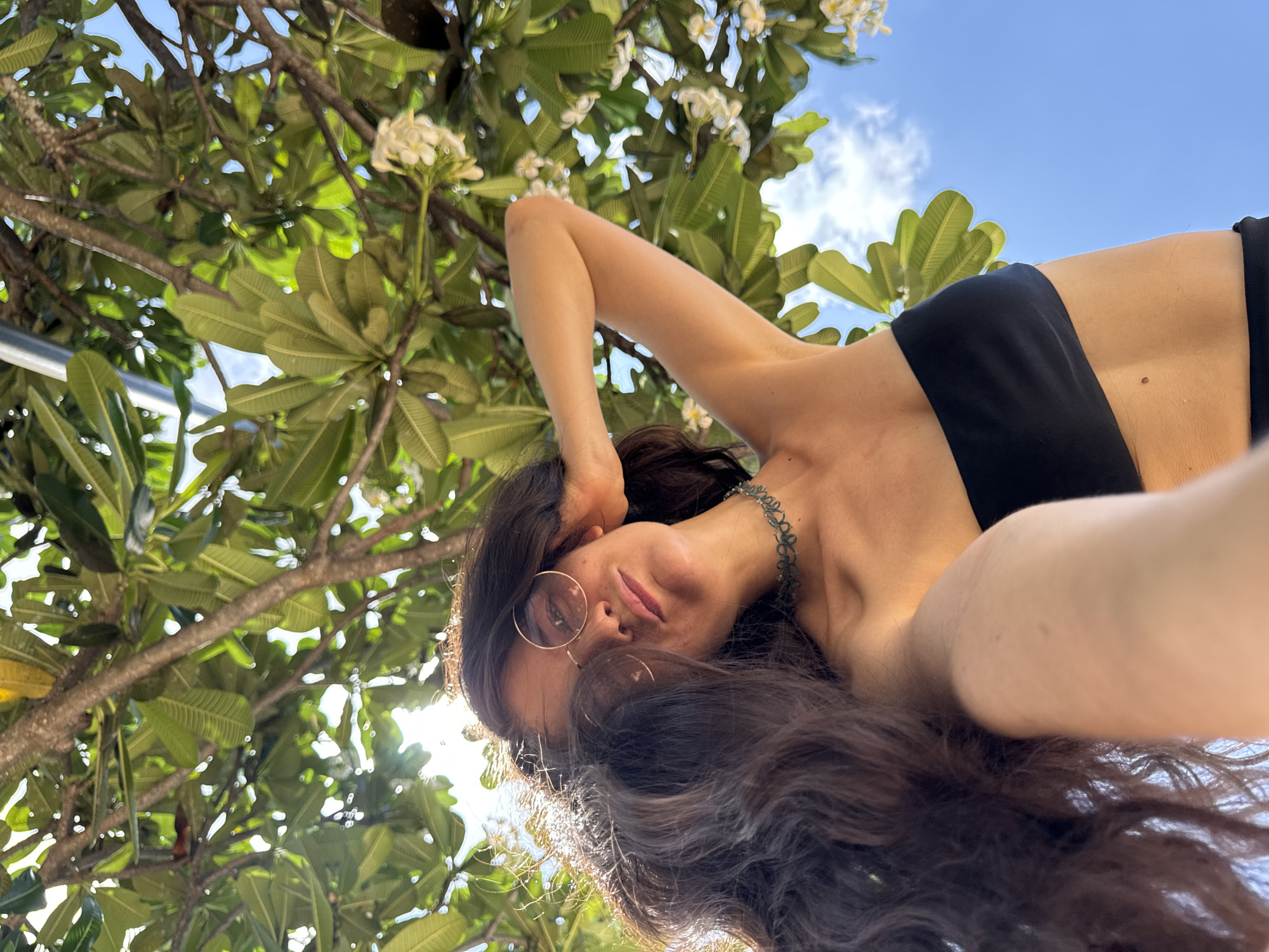 The image depicts a low-angle selfie of a female subject in an outdoor tropical environment in Phu Thuy Ward, Vietnam. The subject, viewed from the chest up, has long, dark, wavy hair that flows over her left shoulder and chest. She is wearing round, light-tinted sunglasses and a black strapless bandeau top. A dark green or teal colored lace choker is visible around her neck. Her left arm is raised, with her hand positioned behind her head. She looks directly into the camera with a neutral expression. The background is a dense canopy of large green leaves and brown branches of a tree, identifiable as Plumeria (frangipani) due to the presence of multiple clusters of small white flowers. Strong sunlight filters through the leaves from the upper left, casting bright highlights. Patches of clear blue sky with minimal white clouds are visible through the foliage, primarily on the right side of the frame.