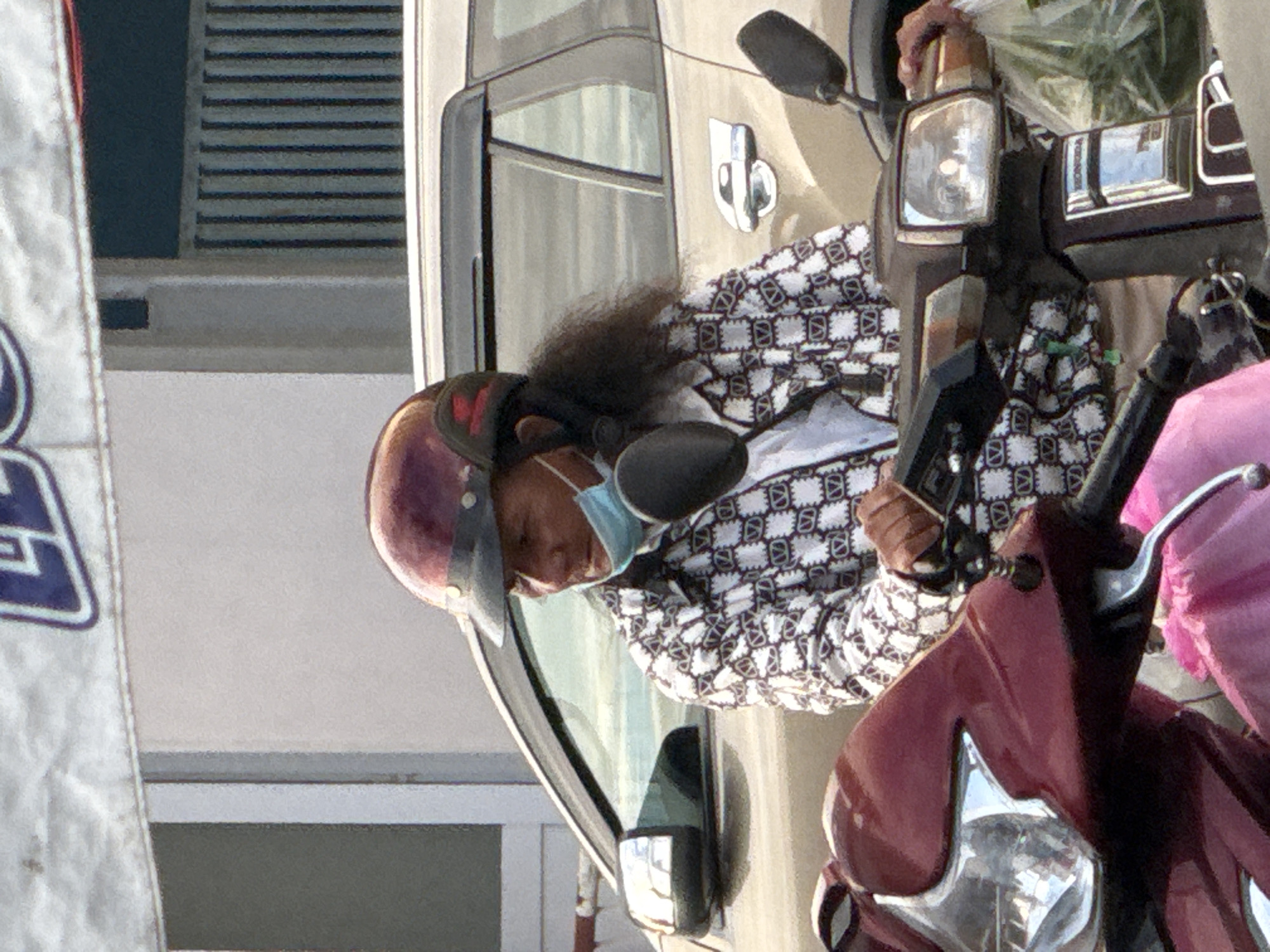 The image shows a person wearing a maroon helmet and a white and black patterned shirt, with a blue medical mask on their face, sitting on a motorcycle. They are looking to the right, with their right hand on the motorcycle's handlebars. A car is visible behind the motorcycle, and a building with vertical blinds is in the background. The scene appears to be outdoors, with natural light. The image is taken in Da Lat, Vietnam.