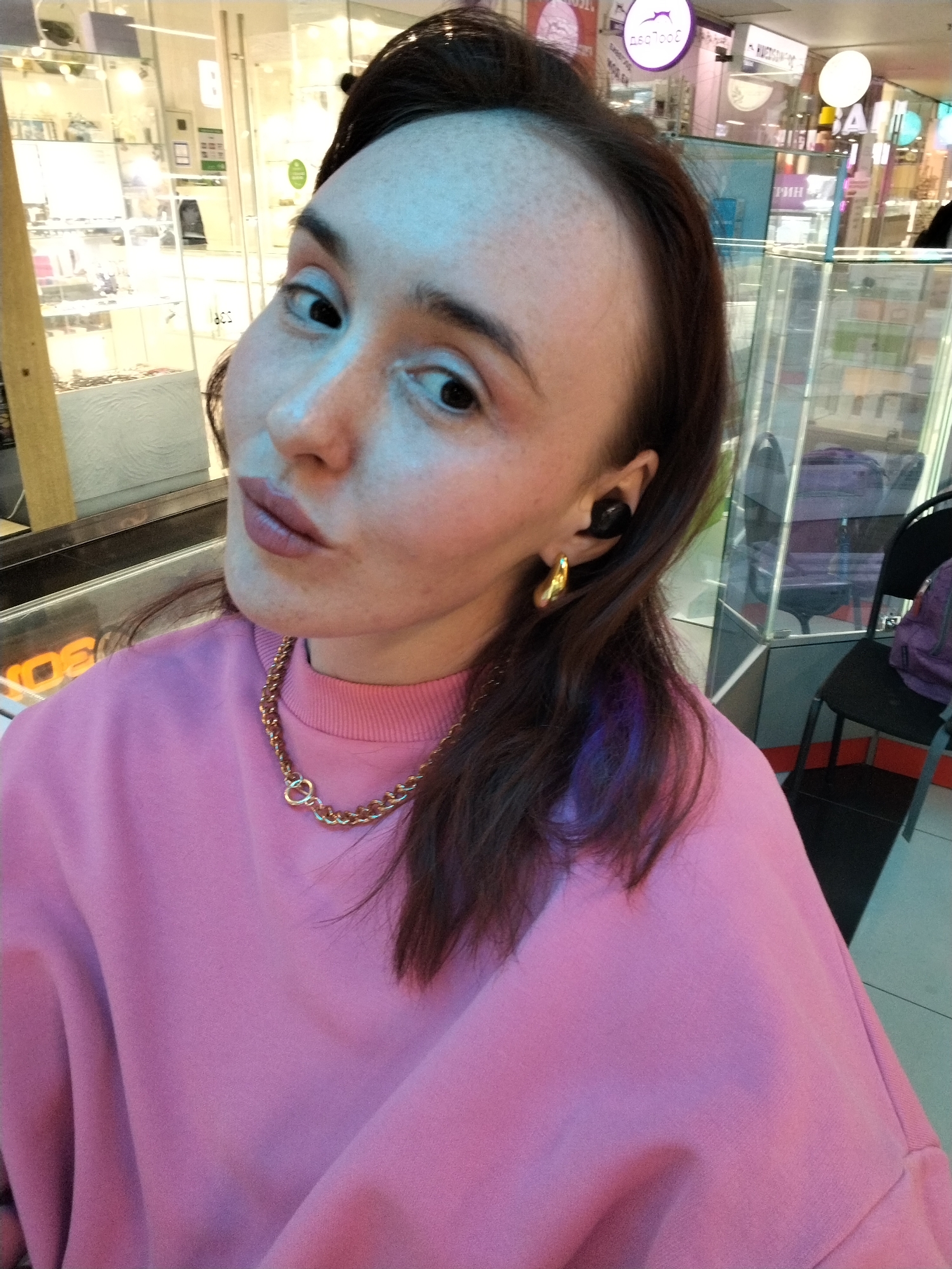 A woman with brown hair and freckles is looking at the camera with puckered lips. She is wearing a pink sweatshirt and a gold chain necklace and earrings. She has wireless earbuds in one ear.
The background shows a brightly lit indoor location, likely a shopping mall. There are glass display cases and storefronts visible. A sign reading "seafood" in Cyrillic characters indicates a nearby restaurant. The city is Izhevsk, Russia, based on the Cyrillic alphabet. A black chair is located to the right.