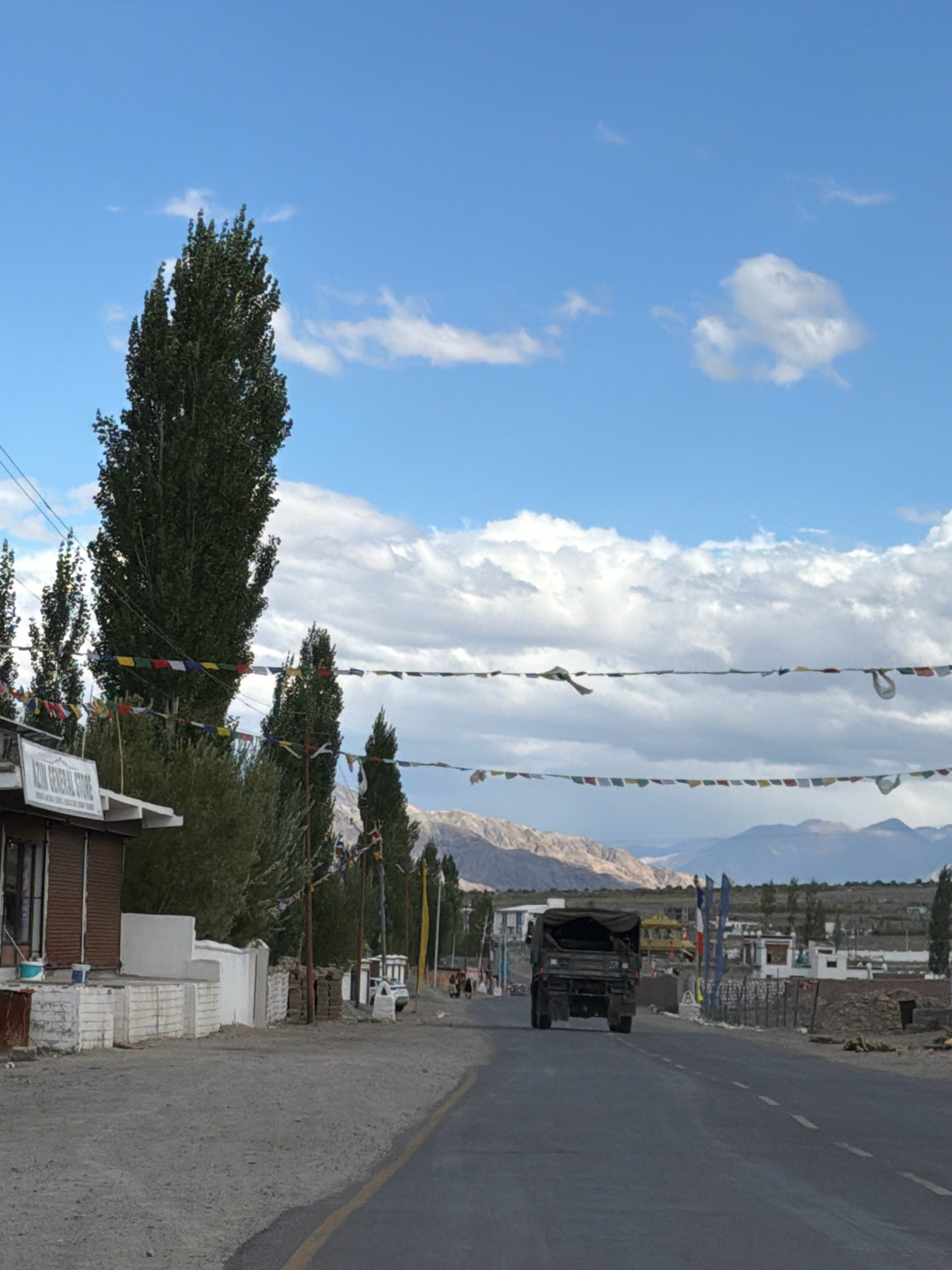 The image depicts a street scene in Yuluk, India, under a partly cloudy blue sky. In the foreground, a paved road with a yellow line marking stretches into the distance. A military truck with a canvas cover is seen driving away from the camera on the right side of the road. To the left of the road, a building with a sign reading "AZIM GENERAL STORE" is visible. Adjacent to the store are white concrete structures and a pile of bricks. Tall, slender trees with dark green foliage line the street, with prayer flags strung between them. In the background, a range of light-colored mountains can be seen under the sky. A few people are walking in the distance on the right side of the road.