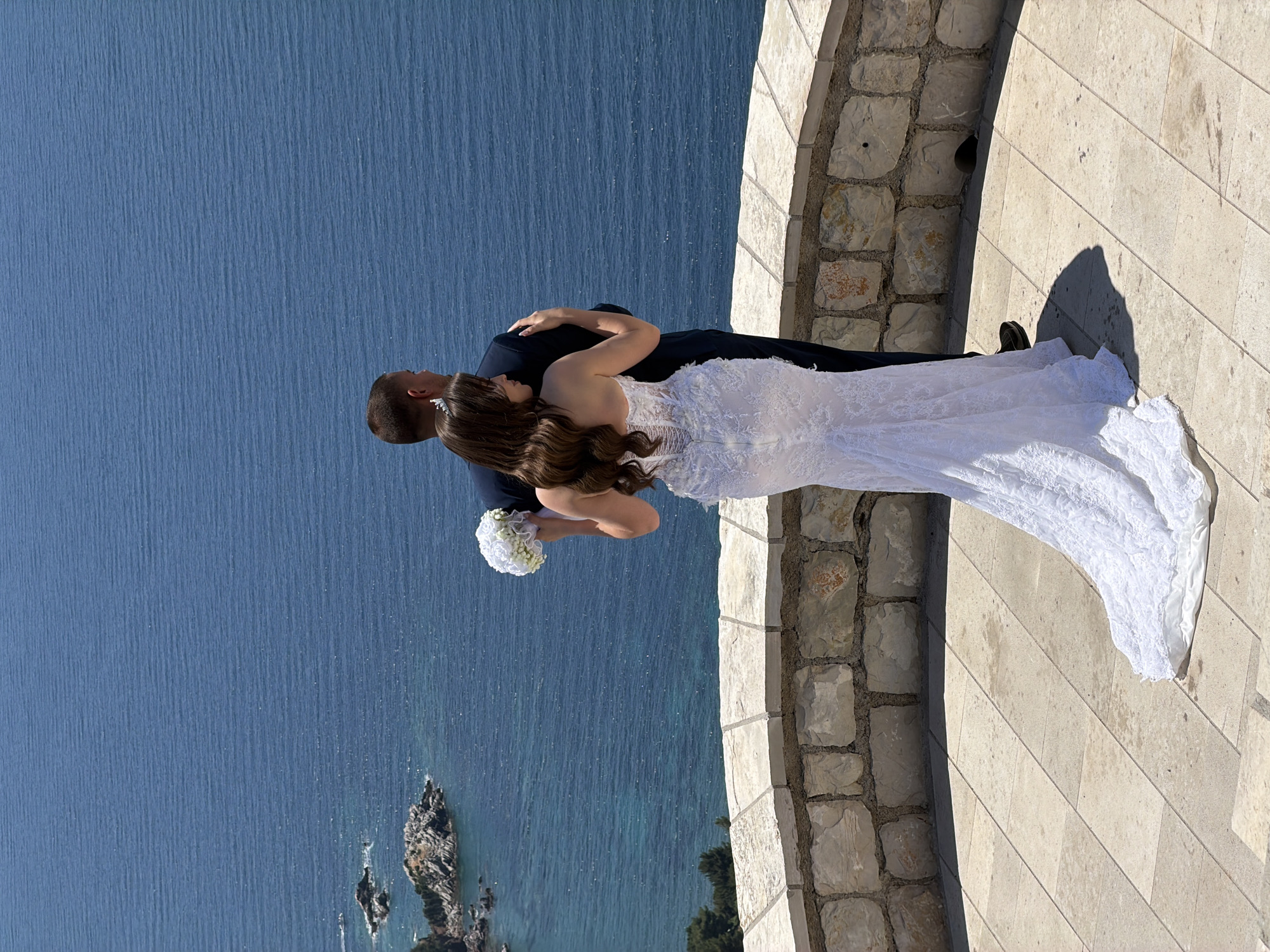 A wedding couple stands on a stone-paved terrace overlooking a blue sea with rocky islands. The woman, wearing a white lace wedding dress and a tiara, has her arm around the man, who is wearing a dark suit. She holds a white bouquet. The sea stretches to the horizon under a clear sky. The terrace is bordered by a low stone wall.