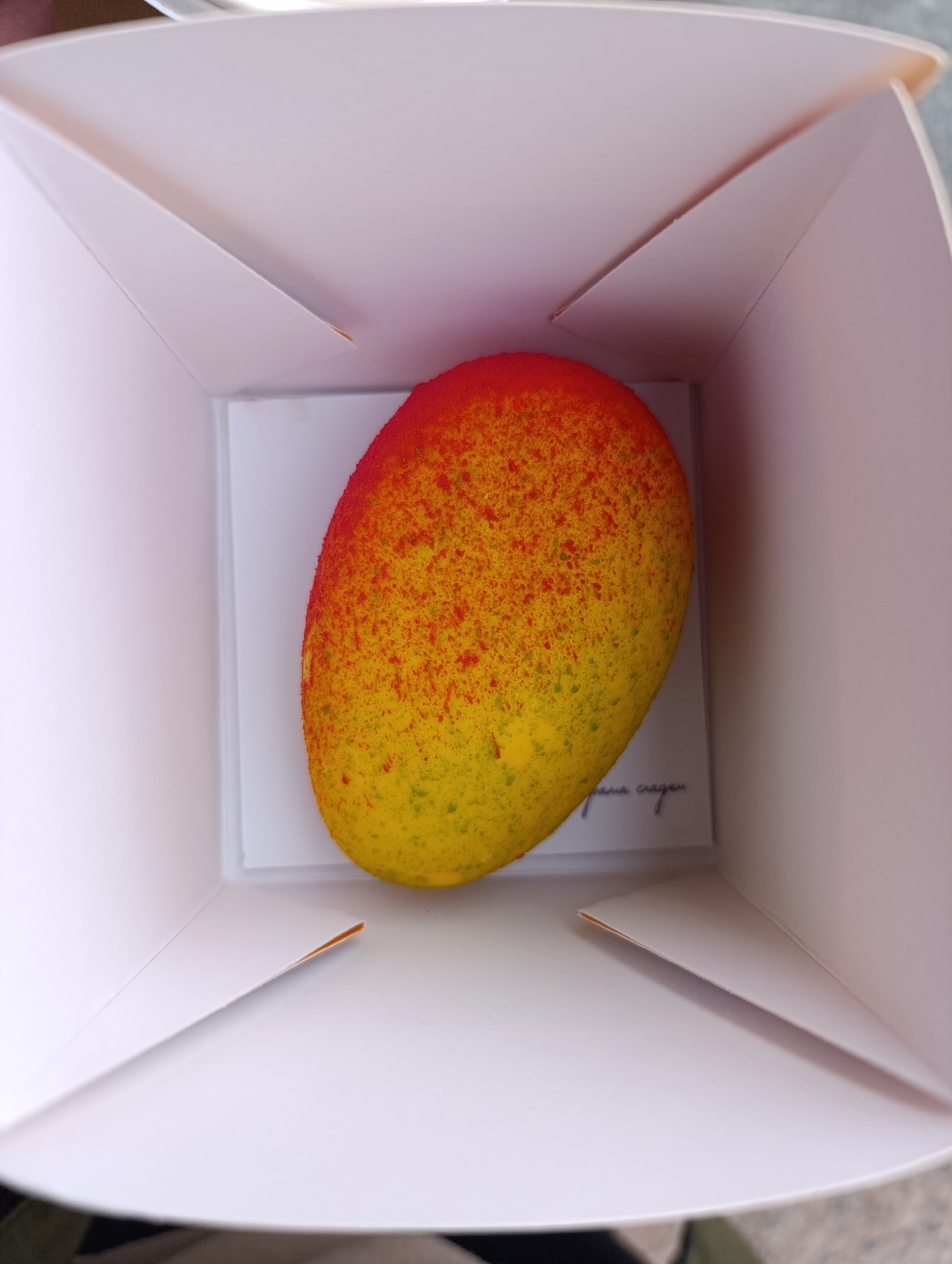 A high-angle, close-up shot shows a cake designed to resemble a mango, contained within a small, white, square box. The cake has a smooth, oval shape, with a gradient of yellow at the base transitioning to red at the top. Small speckles of red are scattered throughout the yellow portion. The cake sits atop a white paper note with faint writing in a cursive-like script. The box is a simple, unfolded design with four triangular flaps forming the corners. The background is out of focus. The image was taken in Sofia, Bulgaria.