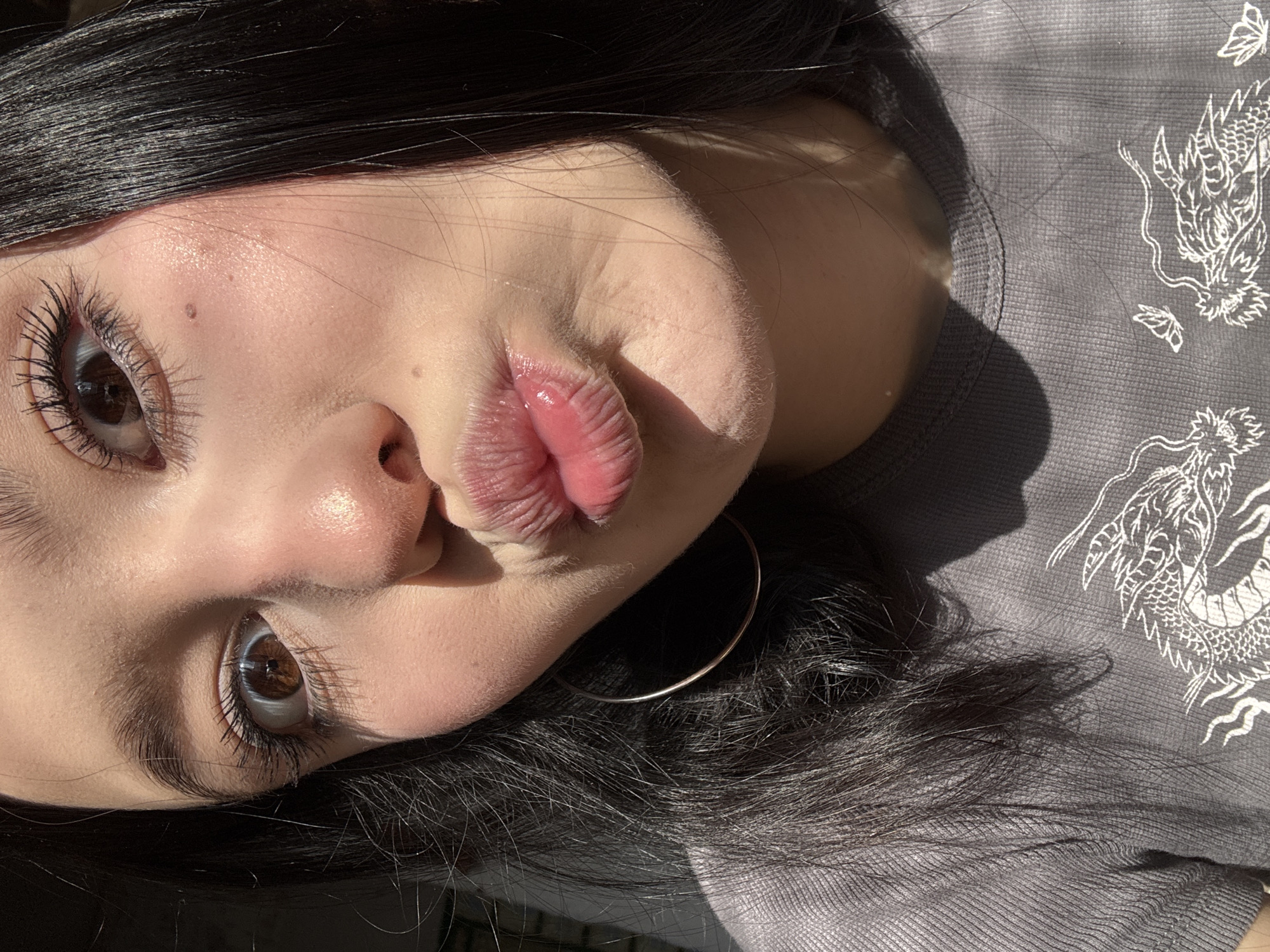 A close-up, angled shot of a person's face with dark hair. The person is pouting their lips towards the camera. The skin is fair with visible shine and texture. The left eye is fully visible, brown in color, with long, dark lashes. A mole is present on the left cheek. A hoop earring is visible near the ear. The person is wearing a gray shirt with a white dragon design on the right side. Lighting creates shadows on the face and shirt. The location is undetermined and there is no information to indicate that it is in the city of Фоки, Russia.