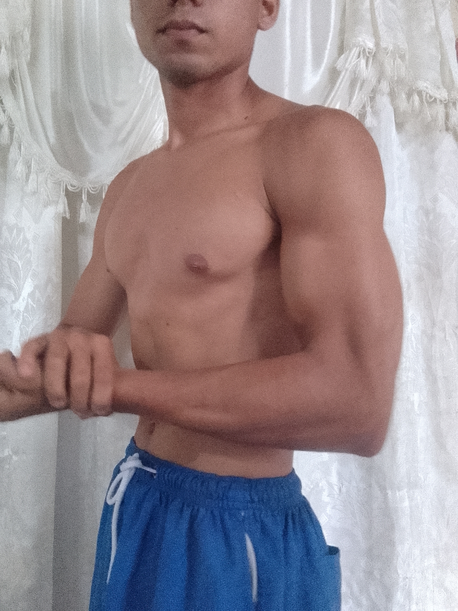 A light-skinned, topless male is posing indoors. He is wearing blue shorts with a white drawstring. The background is a white, textured curtain. The location of Juan Griego, Venezuela is not apparent in the image.
