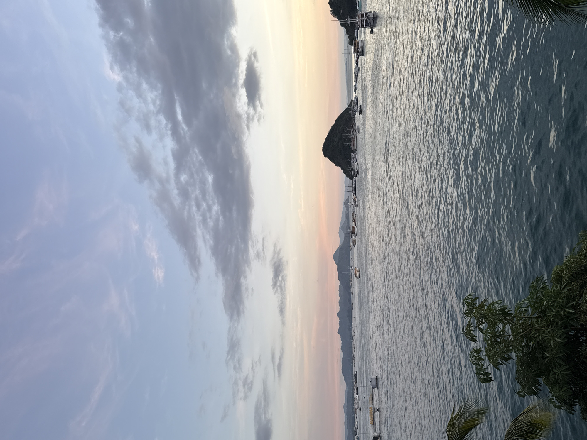 The image shows a coastal scene at sunset in Labuan Bajo, Indonesia. The sky is a gradient of light blue, pink, and yellow, with some dark clouds on the left side. A body of water fills the lower half of the frame, with ripples reflecting the light. There are small islands in the distance and several boats anchored in the water. Some foliage is visible at the bottom right corner.