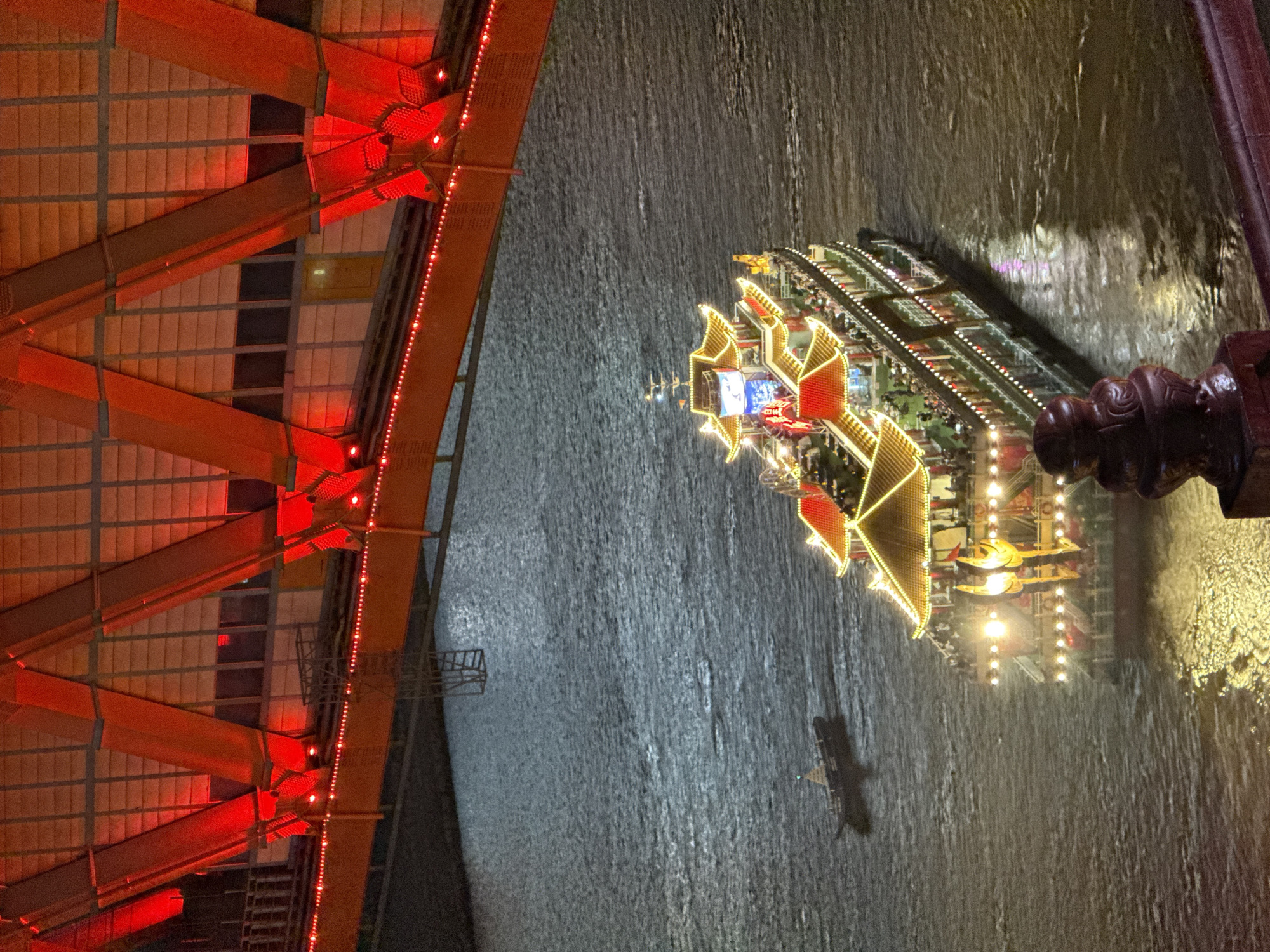 The image shows a section of the Hongyadong complex in the Yuzhong District of Chongqing, China, at night. The complex is lit up with red and gold lights. A decorated boat floats on the river adjacent to the structure.