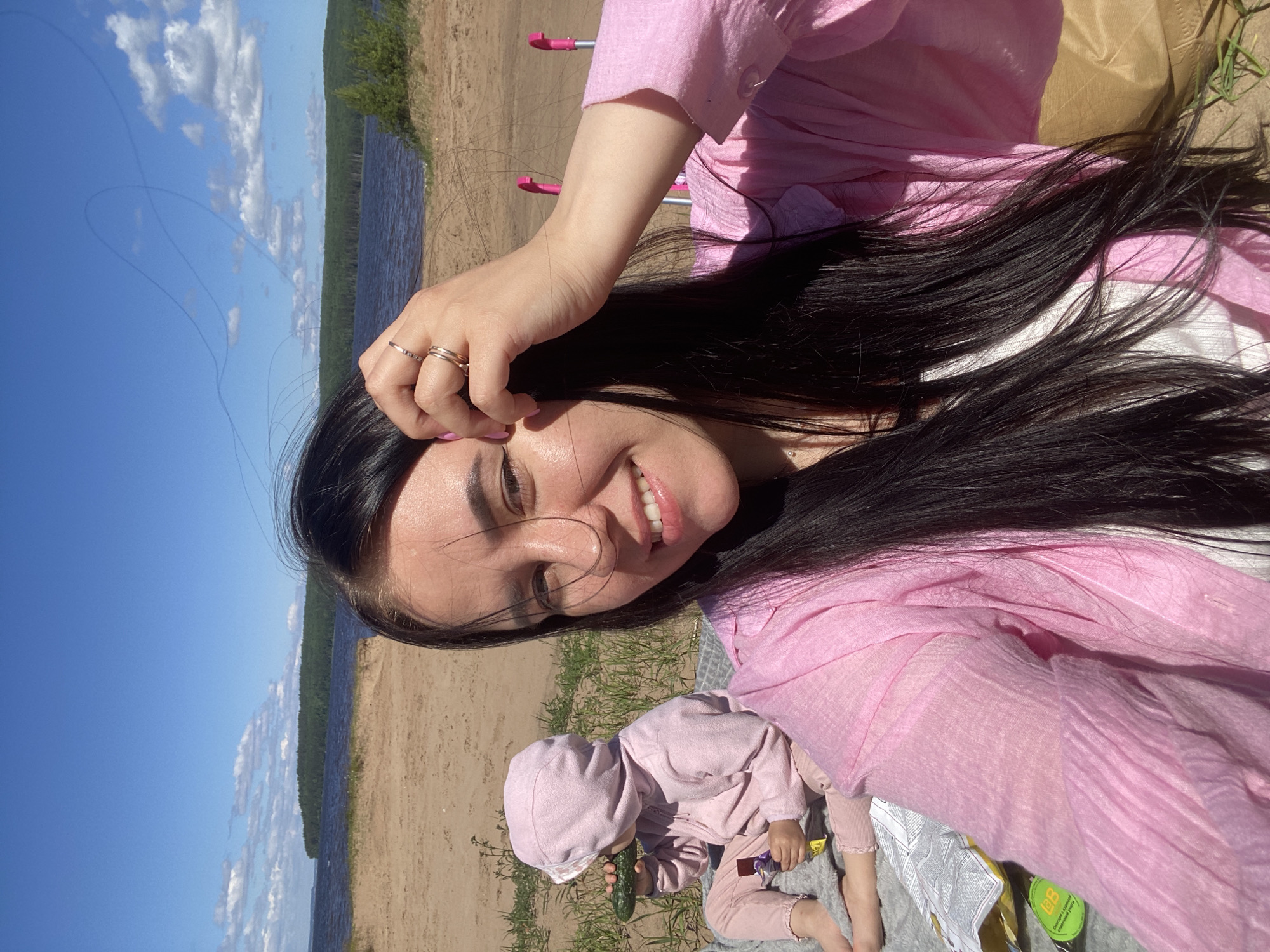 The image shows a woman and a toddler on a sandy beach next to a body of water. The woman is lying down, holding her hand to her head, and smiling at the camera. She is wearing a pink top. The toddler is wearing a pink hooded outfit and is eating a cucumber. There are some scattered green plants on the sand. In the background, there is a body of water, a sandy shore, and a tree line under a blue sky with white clouds. The lighting suggests it is daytime, likely sunny. The context provided indicates the location is Chaikovsky, Russia. There is some visible text on a packet in the foreground, but it is not clearly legible. The overall mood appears relaxed and happy, suggesting a day out at the beach.