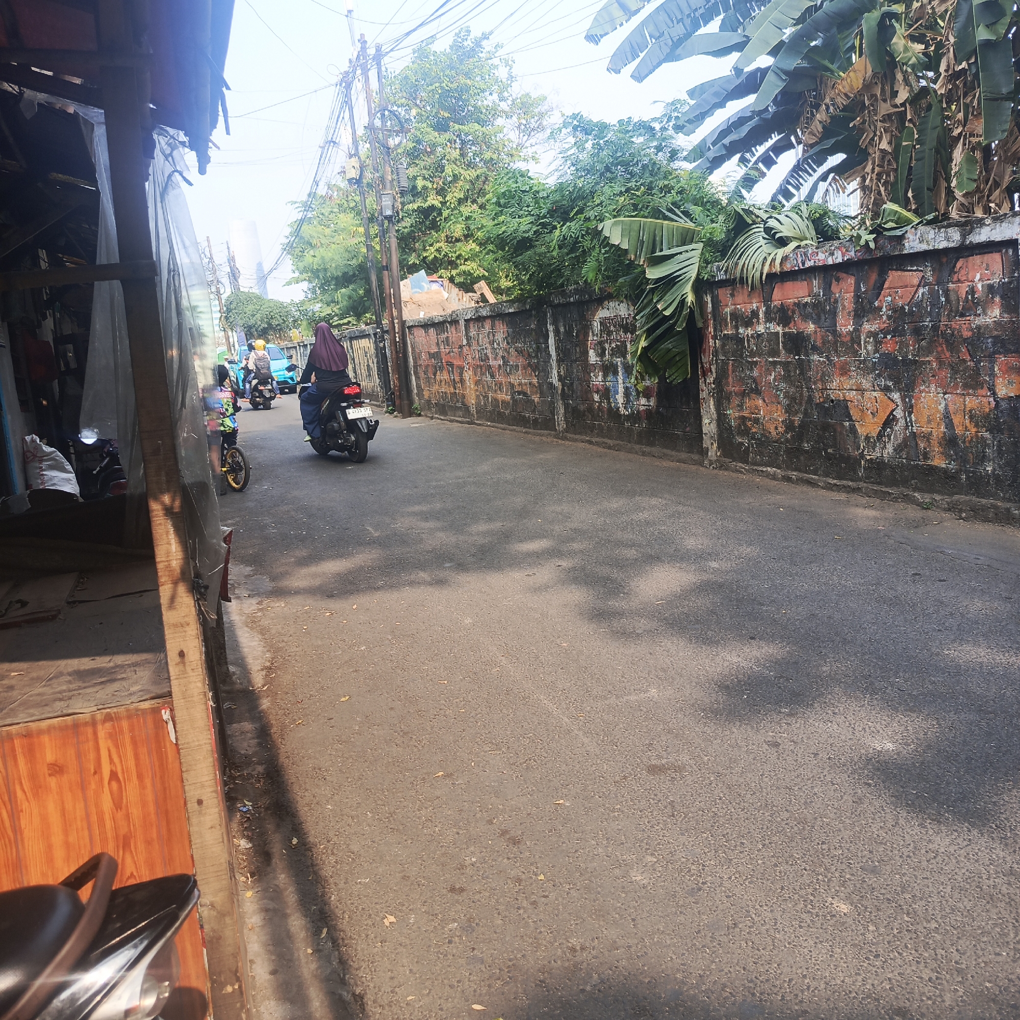 The image depicts a street scene in South Jakarta, Indonesia, on a bright, sunny day. The road surface is asphalt, with visible cracks and some debris. To the left, a wooden structure with a corrugated plastic roof and translucent sheeting suggests a temporary or open-air stall or structure, possibly for vendors. Some items, including white bags, are visible within this structure. A motorcycle is parked near this structure.
The street is lined with a brick wall on the right side, which appears weathered with peeling paint and graffiti in muted colors. Banana leaves and other greenery are visible above the wall, indicating the presence of trees and vegetation. Power lines and poles are prominent against the bright sky, a common feature in urban Indonesian environments.
Several individuals are on motorcycles on the road. One motorcycle, closer to the foreground and on the right side of the road, is ridden by a person wearing a dark hijab and blue pants. Another motorcycle is further down the road, with a rider wearing a yellow helmet. A blue car is also visible in the distance, suggesting light traffic. The overall impression is a busy, functional street in a tropical urban setting.