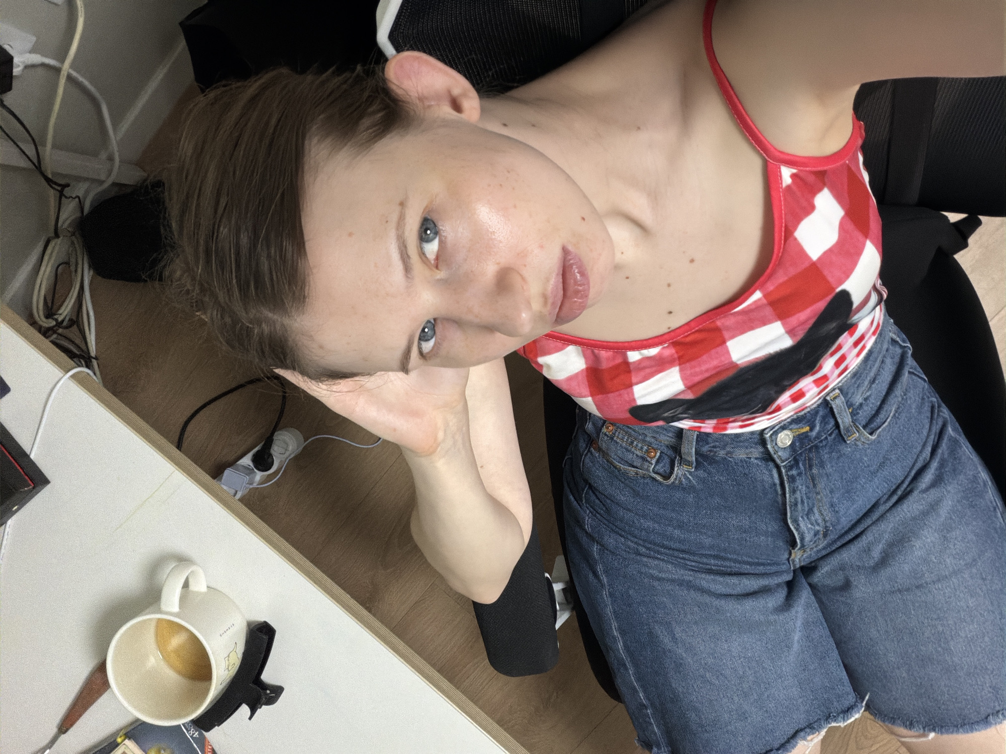 The image captures a young woman sitting at a desk, possibly in an office or study environment. She is angled away from the camera, with her head turned to look directly at it. Her right arm is bent and resting behind her head, and her legs are crossed, with her left foot resting on the armrest of her chair. She is wearing a red and white checkered tank top and denim shorts. On the desk beside her is a white mug with a yellow liquid inside, likely coffee or tea, and a black clip attached to the mug. Various cords and electronic devices are visible on the floor and desk, suggesting a workspace. The overall lighting suggests an indoor setting, possibly during the day. The user does not have permission to use the location information as no location is present in the image.