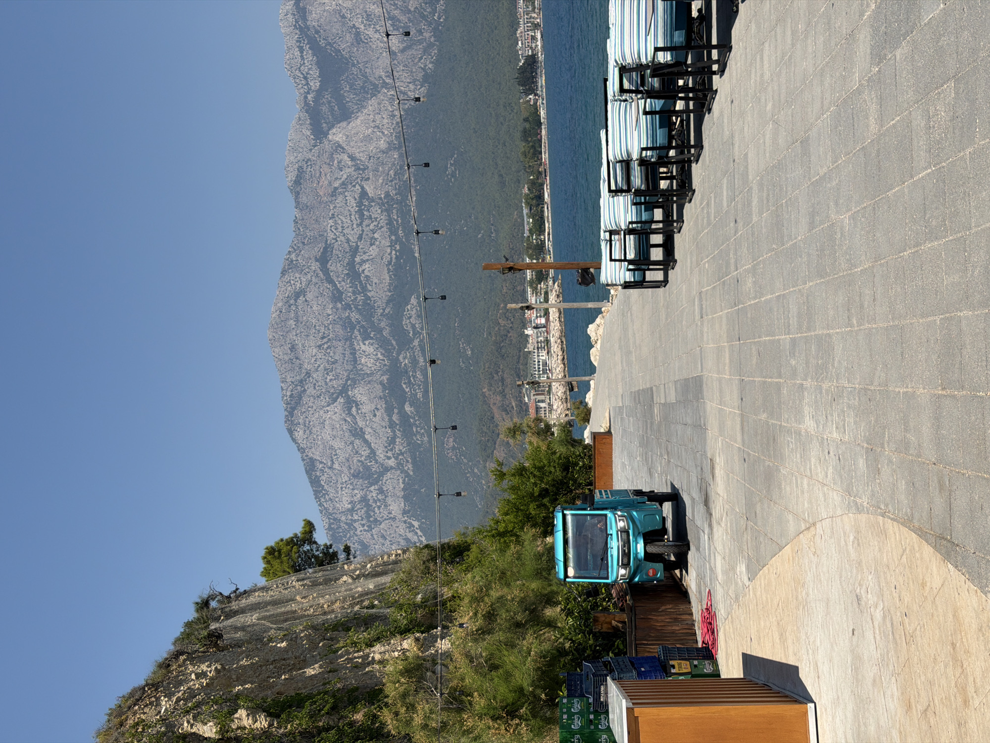 A bright, sunny day is depicted in this image, featuring a coastal landscape. The scene is oriented vertically. In the foreground, a light blue utility vehicle with a small cab is parked on a paved area. Beside it, stacked crates are visible, some appearing to be for beverages. Behind the vehicle, lush green foliage and a rocky hillside covered with netting are present. A string of outdoor lights stretches across the frame.
In the midground, the blue sea meets a coastline lined with buildings and green trees. The background is dominated by a large, grey, rocky mountain range under a clear blue sky. To the right, a row of empty lounge chairs with blue and white striped cushions are neatly arranged on a paved walkway, suggesting a resort or public seating area.
The time of day appears to be late morning or early afternoon, given the strong sunlight casting shadows. The overall mood is peaceful and scenic, with no visible human activity beyond what might be inside the utility vehicle. No discernible text is visible in the image. The location suggests a Mediterranean or similar climate, with the juxtaposition of mountains, sea, and manicured seating areas.