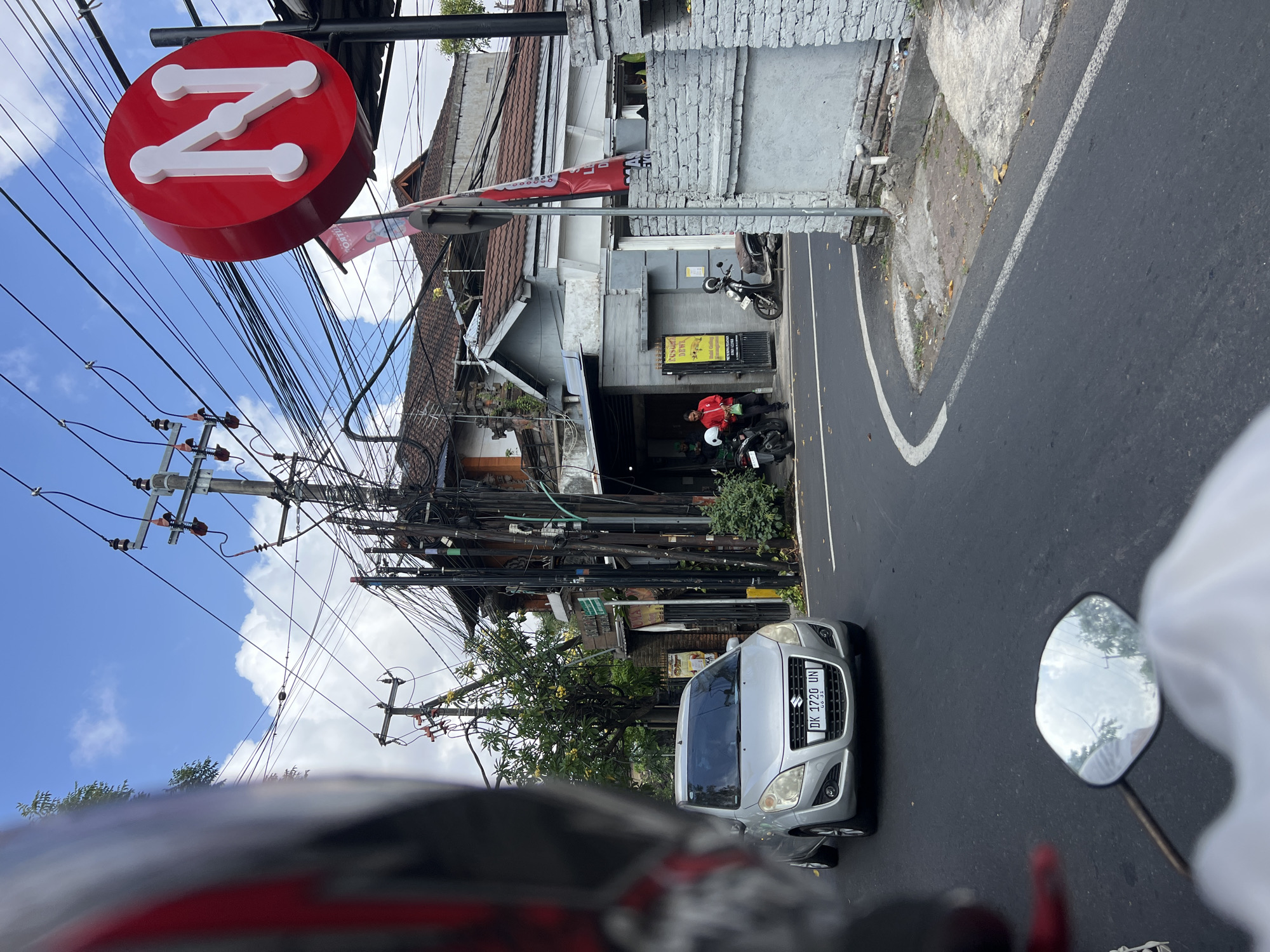 A silver Suzuki hatchback with license plate DK 1720 UN is driving on a paved road in Denpasar, Indonesia. The car is in the foreground, slightly to the left of center. Behind the car, there are several buildings with tiled roofs and a prominent red sign with a white letter "N" on it. Numerous electrical wires are strung between poles, obscuring the blue sky and white clouds. Two individuals, one wearing a red jacket and the other in a white shirt, are standing near motorcycles outside one of the buildings. The road is marked with white lines, and there is some debris on the side of the road. The image appears to be taken from the perspective of someone on a motorcycle, with a portion of a helmet visible in the lower left corner.