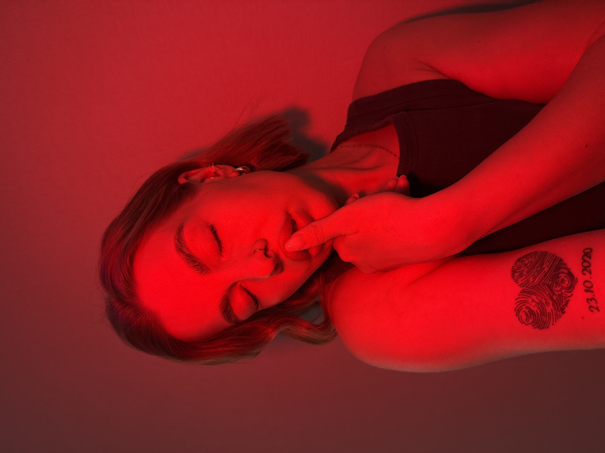 The image shows a person lying down with their eyes closed, illuminated by a red light. Their right hand is touching their face near their mouth. The person has a tattoo on their left forearm consisting of two overlapping fingerprint hearts and the date "23.10.2020". The image does not contain any information about the location or the city of Chaikovsky, Russia.