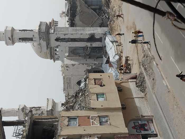 In Khan Yunis, Palestinian Territory, an urban street scene displays extensive structural damage under a hazy sky. A large mosque with a light-colored dome and two minarets is prominent in the background, exhibiting severe damage to its facade, shattered windows, and partial collapse on its minarets and lower sections. To the left of the mosque, a multi-story building is significantly destroyed, with its upper floors reduced to rubble and exposed rebar, while lower levels show numerous impact marks and broken windows. A "Squid Game" poster is visible on the ground floor of this building. In the midground, an open, sandy area hosts several white, tarp-covered structures, consistent with temporary shelters. Adjacent to these, a donkey-drawn cart loaded with items, likely produce, is stationary with a person standing nearby. The foreground features a paved road with a visible white dividing line. Approximately ten individuals are present: a group of five (two adults, three children) walks on the road from right to left, another pair walks further left on the road, and additional individuals are observed walking in the sandy areas near the tents and damaged structures. Piles of debris and construction rubble line the edges of the road and damaged buildings. The image is captured from an elevated perspective, indicated by an out-of-focus metal railing in the lower right foreground.