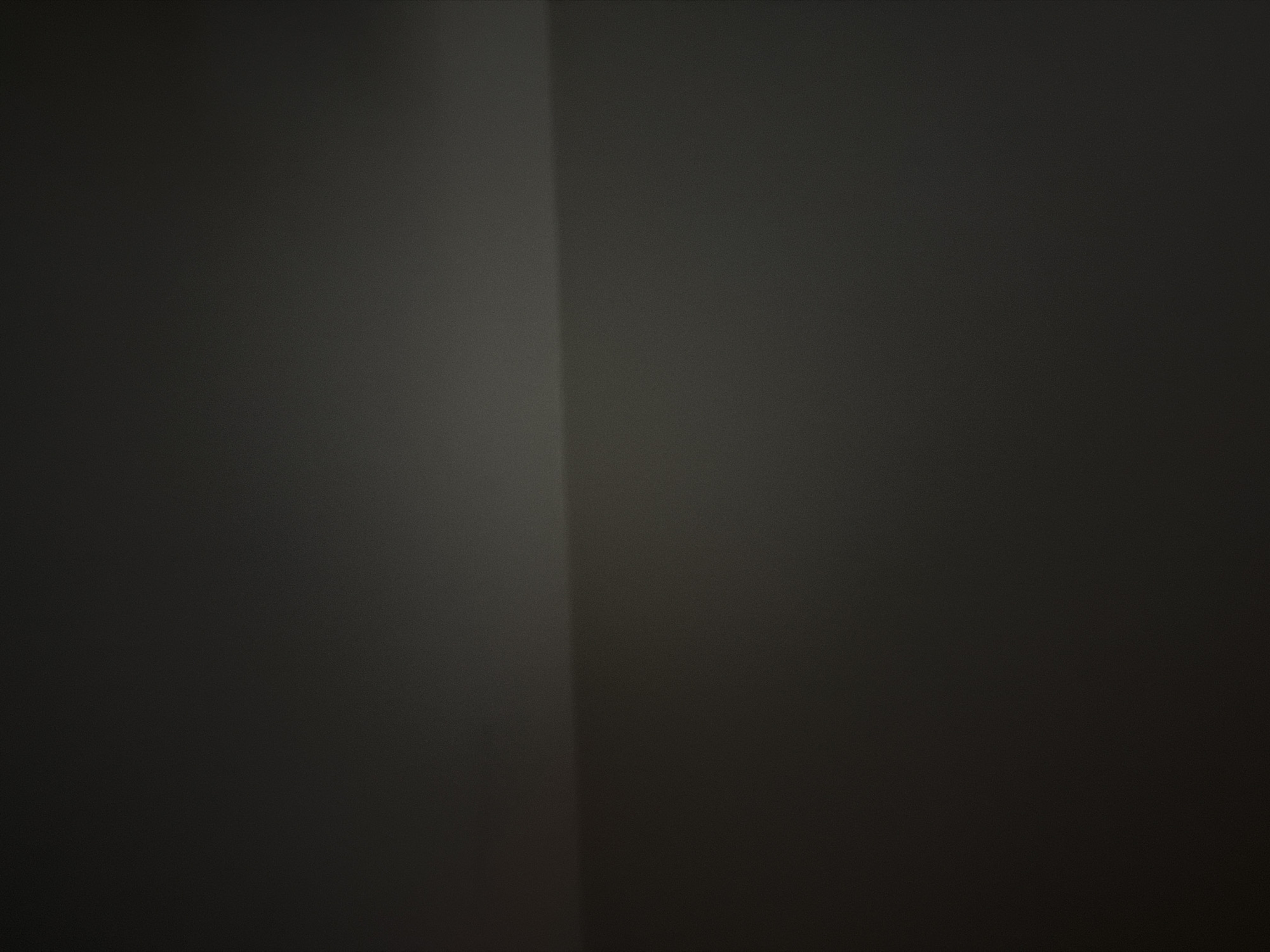 The image is a dark, abstract photograph with a horizontal line dividing the frame. The top portion is a gradient of dark gray to black, with a subtle highlight on the right edge. The bottom portion is a darker shade of gray, also with a gradient effect. There are no discernible objects, people, or actions. The environment appears to be an interior space with low lighting, possibly a wall or a surface. There is no identifiable location in The Hague, Netherlands.