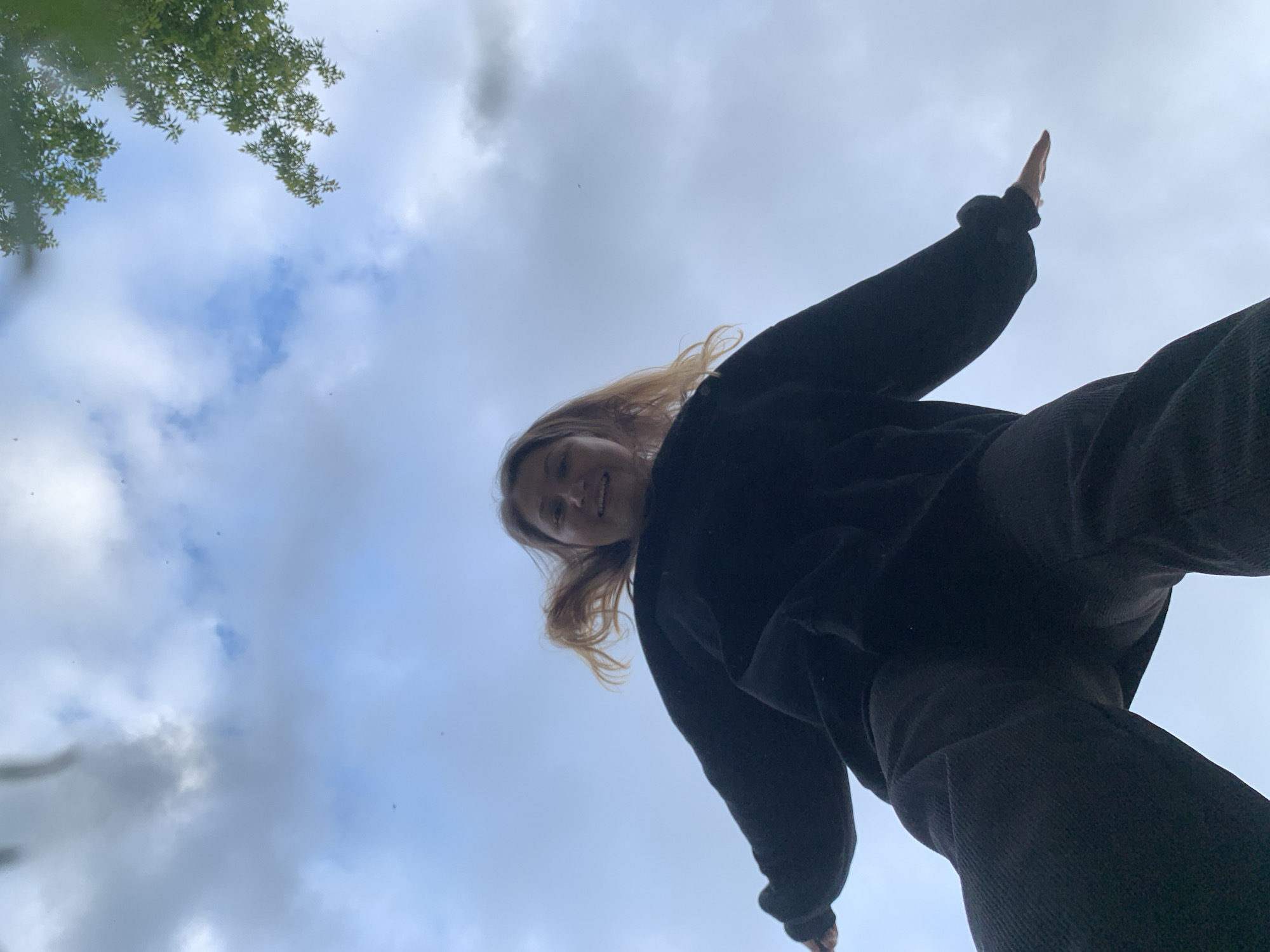 A low-angle shot captures a person in mid-air against a cloudy sky. The person, viewed from below, is wearing a black sweatshirt and dark pants. Their arms are outstretched, with the right arm extended to the side and the left arm bent at the elbow. Their hair is light brown and appears windswept. A small portion of green foliage is visible in the upper right corner of the frame. The overall scene suggests an outdoor activity, possibly a jump or a playful pose. There is no discernible urban environment or specific location within Chaikovsky, Russia visible in the image.