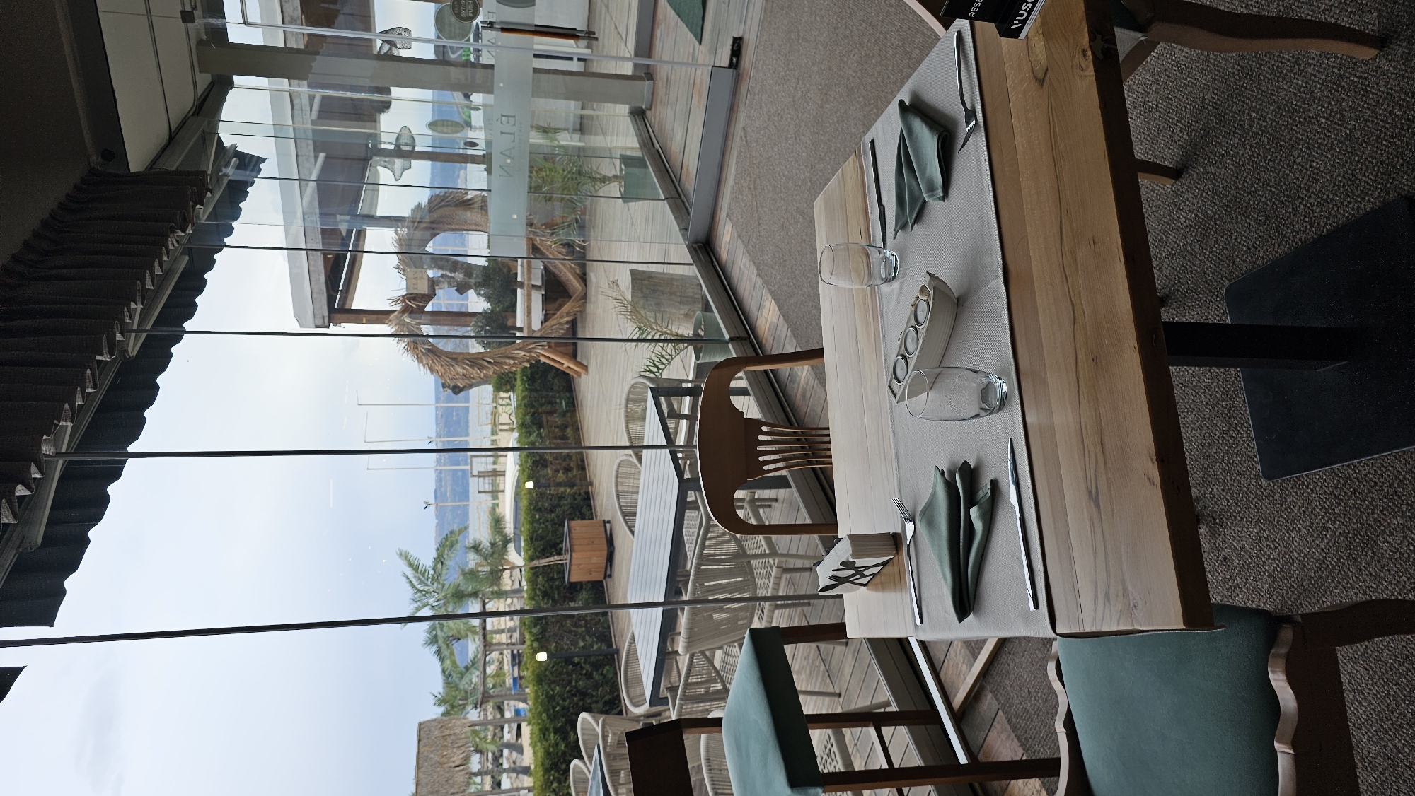 The image depicts a restaurant interior with a view of the beach and sea. The setting appears to be sunny, likely in Varna, Bulgaria, given the presence of beachside palm trees and the sea.
A rectangular wooden table is the central focus. Table is set with a light gray runner, napkins, cutlery, glassware, and a unique salt/pepper shaker or candle holder. Chairs with wooden frames and blue-green fabric seats are placed around the table.
Outside the restaurant's glass windows is a beach area with palm trees, other beach structures, and a body of water. The outdoor furniture is visible.
There are no people present in the photograph. The scene focuses on the ambiance of a seaside restaurant setup.