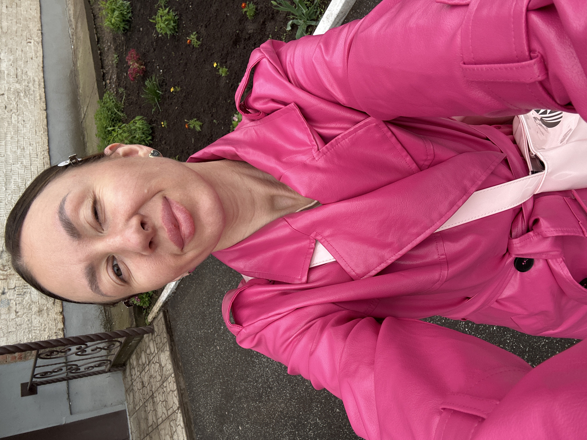 A person is wearing a bright pink leather trench coat. The person is outdoors. There is a flower bed with various plants behind the person. There appears to be a wrought iron railing next to a building, which suggests an urban location. Without more information, it is not possible to confirm this location to be in Votkinsk, Russia.