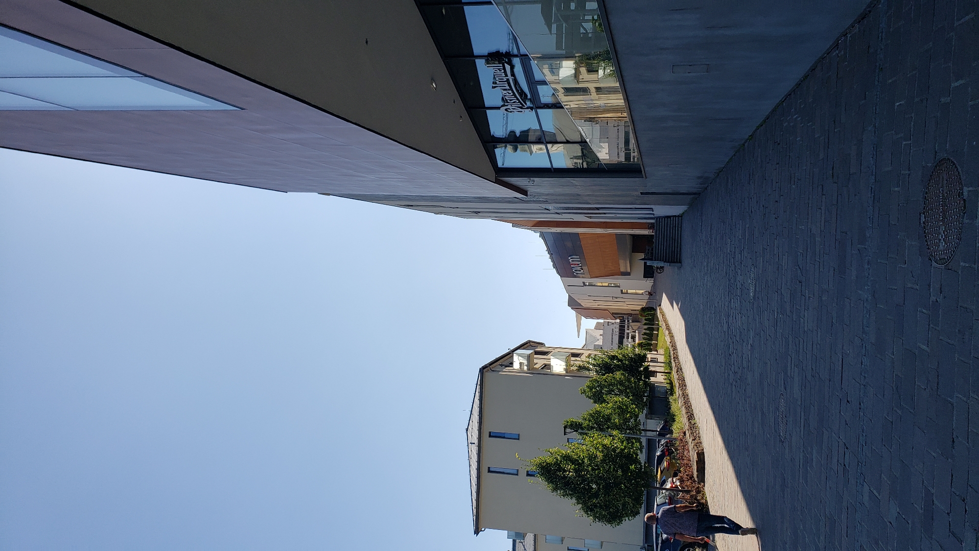 The image depicts a street scene in the city of Prešov, Slovakia, likely in a modern commercial or mixed-use area. On the right, a large building with a grey facade and reflective windows dominates the foreground. The windows of this building display a reflection of a church tower and other buildings. Text on the window reads "Pilsner Urquell". To the left, a pedestrian walkway paved with dark grey stones leads away from the viewer, bordered by trees and a lighter colored building. Further down the street, a building with a large "novum" sign is visible. A person is walking away from the camera on the left side of the walkway, wearing a blue shirt and dark pants. The sky is clear and blue, indicating a sunny day. The overall impression is one of urban development with contemporary architecture.