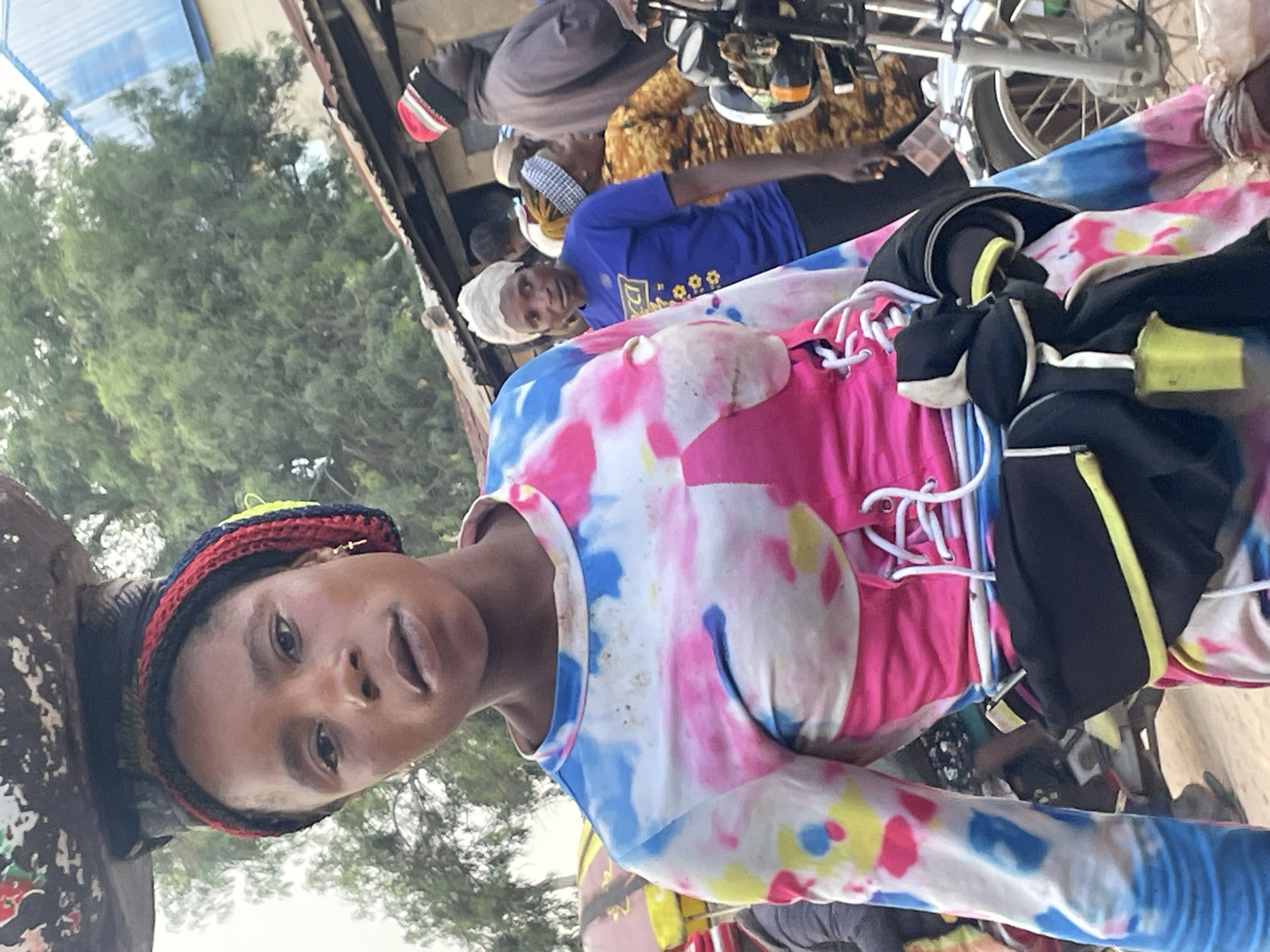 A young Nigerian woman stands in a marketplace in Wang, Nigeria, carrying a large, dark object balanced on her head. She wears a colorful tie-dye shirt with pink and blue accents and a cinched waist secured by white laces. A black fanny pack with a yellow trim is worn around her waist. In the background, other people are visible, along with market stalls and a motorcycle. The environment appears to be an outdoor market with some rudimentary structures.