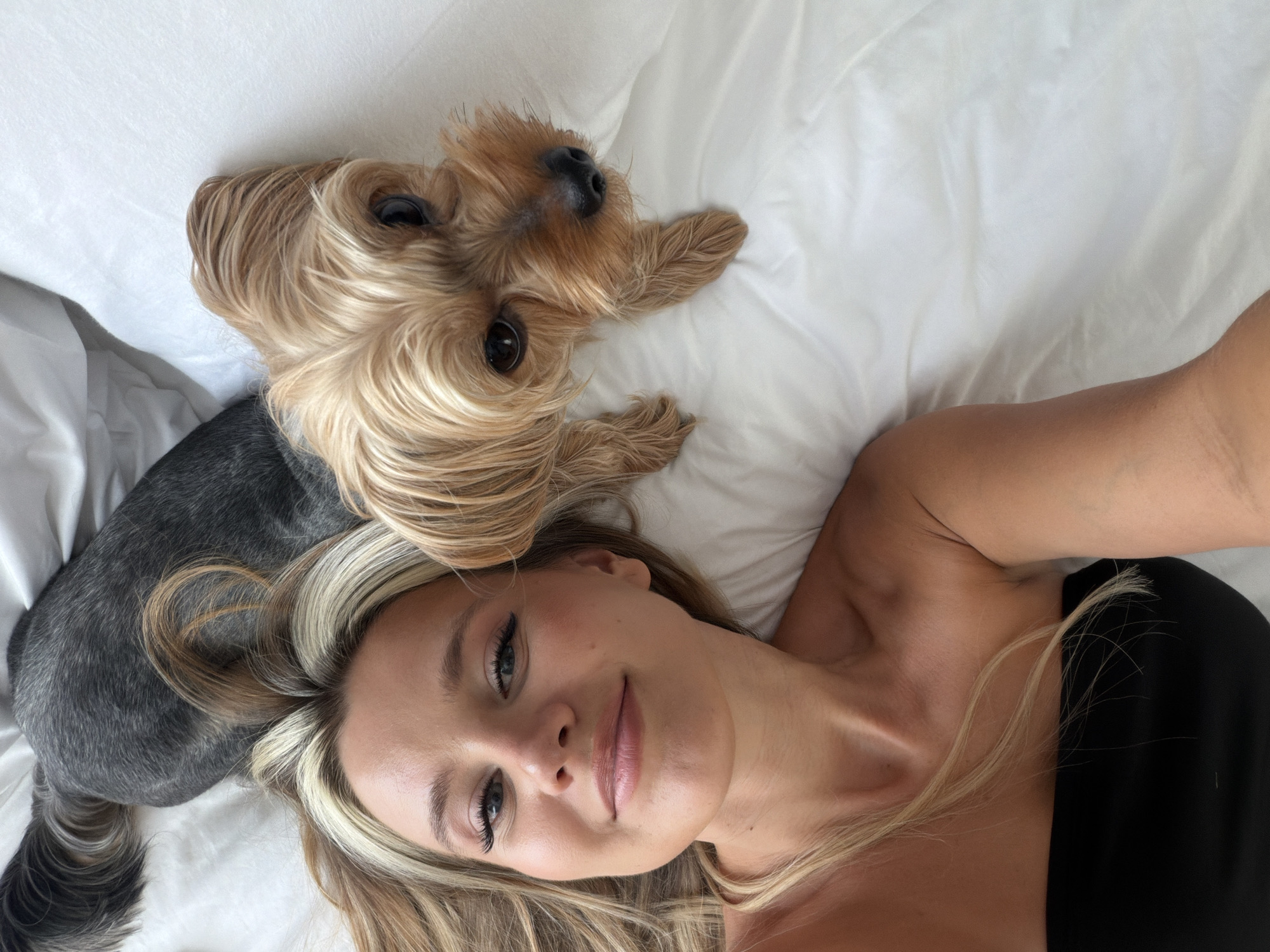 A young woman and a Yorkshire terrier dog lie on a white bed. The woman is in the foreground, facing the camera and smiling. She has blonde hair with streaks of lighter blonde and is wearing a black strapless top. The dog is to her right, looking up at the camera. Its fur is brown and tan. In the background, the back and tail of another dog, possibly a grey schnauzer mix, are visible. The scene is well-lit, suggesting it was taken during the day. The room appears to be a bedroom, with white linens on the bed and a subtle texture of the fabric visible. There is no indication of the city of Thep Krasatti, Thailand.