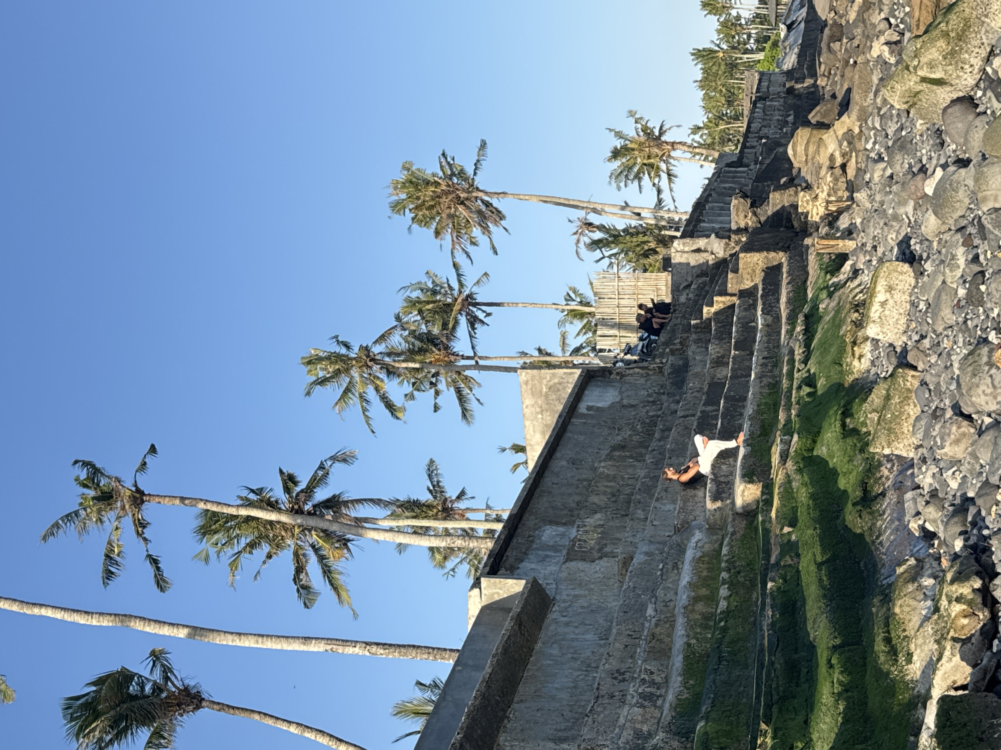 A wide shot shows a person sitting on a stone staircase leading down to a rocky beach, with palm trees and a clear blue sky in the background. The staircase is weathered and covered in green moss in places. The person is wearing white pants and a dark top, sitting with their legs crossed. In the distance, a few other people are gathered near a bamboo fence, and a motorcycle is parked on a higher level of the structure. The sea is not visible, but the presence of the beach suggests a coastal location, potentially in Mengwi, Indonesia, given the abundance of palm trees. The overall scene depicts a relaxed, outdoor setting under bright sunlight.