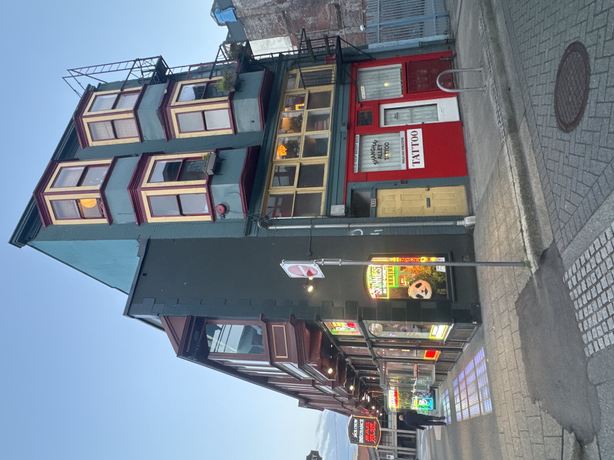 The image shows a street-level view of a three-story building with a teal facade and dark green accents, located in Vancouver, Canada. The building features multiple bay windows with red and yellow trim on the upper floors. Neon signs are visible on the ground floor, including "JACK CHOW INSURANCE" in red, and "SKINZIES IN THE WORLD" in yellow, with an illuminated panda display in the window. One storefront is painted bright red with the words "SHANGHAI ALLEY TATTOO" and "TATTOO" displayed prominently. A yellow door is visible to the left of the tattoo shop, and a white door is to its right. The street is paved with cobblestones, and a sidewalk runs along the front of the building. A single person is walking away from the camera on the sidewalk, casting a reflection from the storefront lights. A metal fire escape extends vertically along the side of the building. The sky is a clear, pale blue, indicating it is likely dusk or early morning. A traffic sign with a red circle and a horizontal bar is mounted on a pole in front of the building.