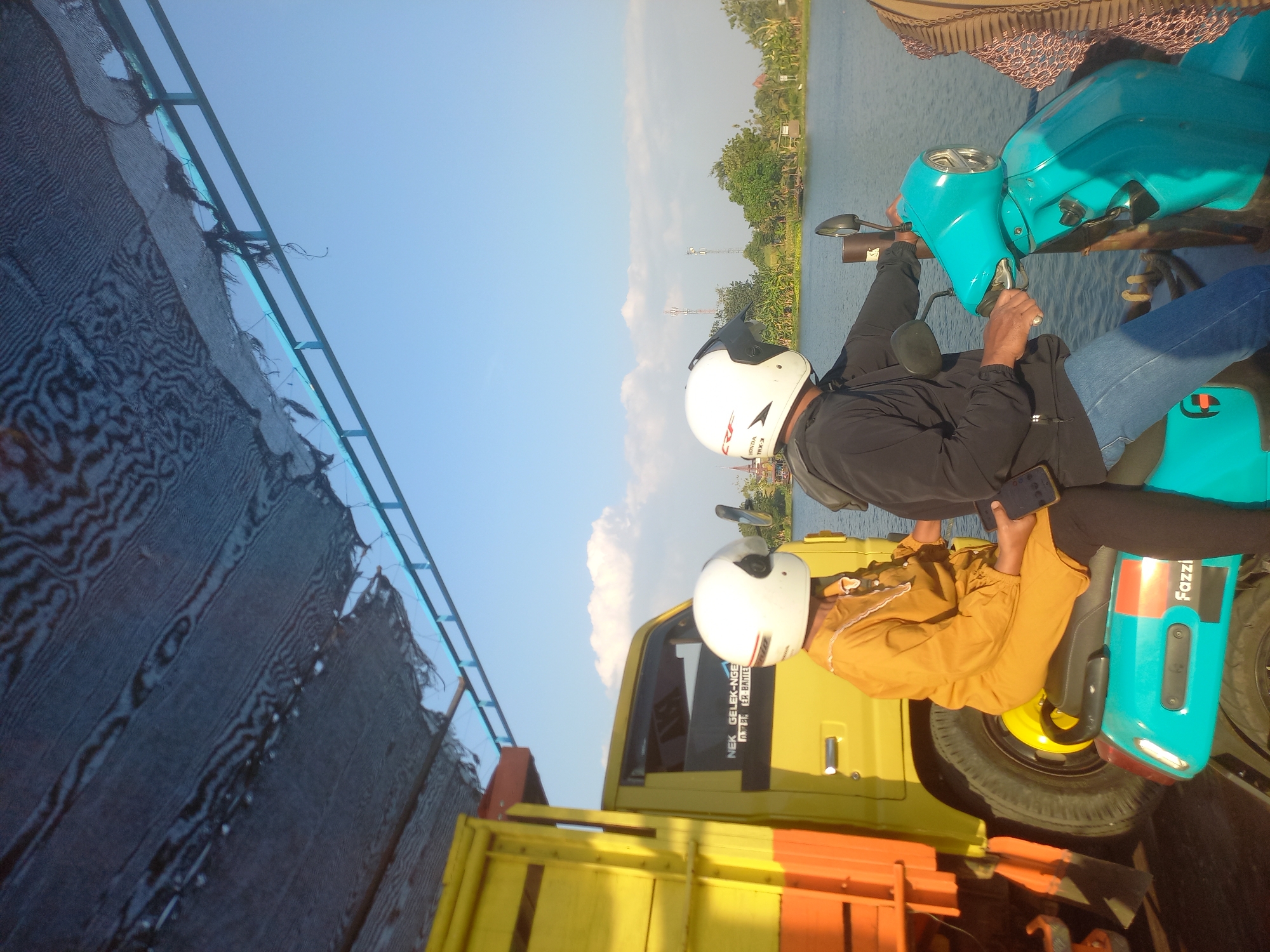 The image shows two people on a blue electric scooter parked next to a yellow truck. The people are wearing helmets, and the scooter is parked near a body of water. The scene appears to be in the city of Munung, Indonesia, as suggested by the text on the truck. The truck has text that reads "NEK GELEK-NGELEK". The sky is clear and blue, with a few clouds. The overall scene suggests a casual outing or travel.