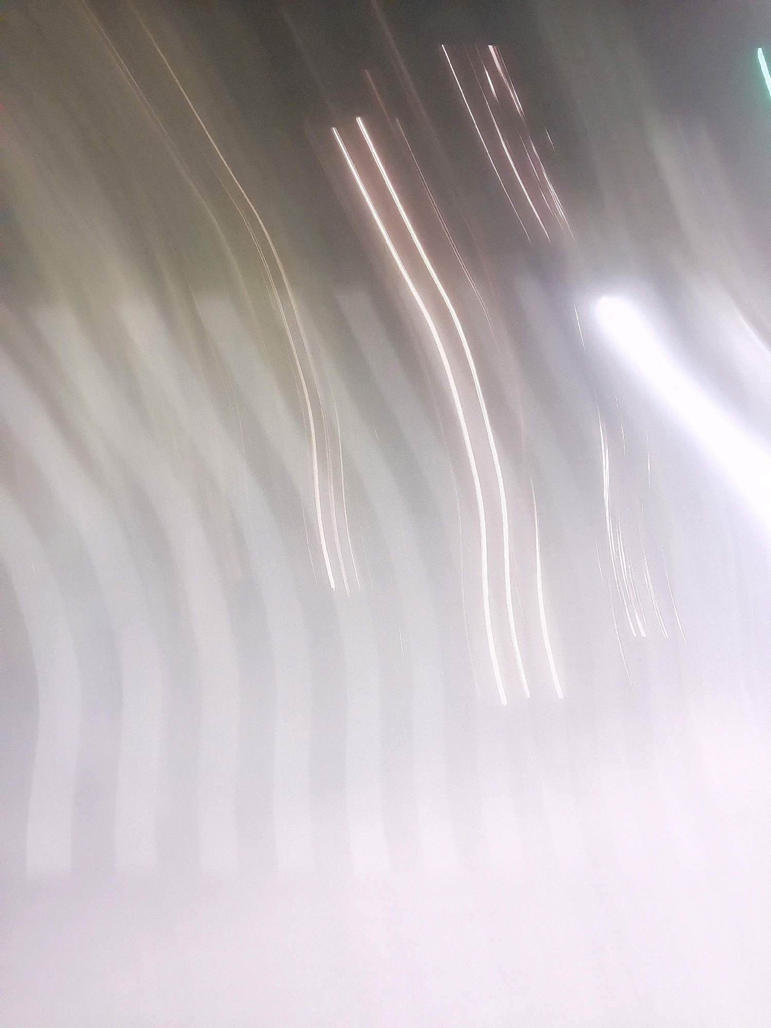 This is an abstract, blurry image with streaks of light against a muted background. The streaks appear to be a result of long exposure photography, capturing the movement of light sources. The colors are predominantly light gray, white, and hints of pale pink and green. There are no discernible people, objects, actions, interactions, or environmental features that would allow for a description of a specific location, including Sleman, Indonesia. The image is purely abstract light trails.