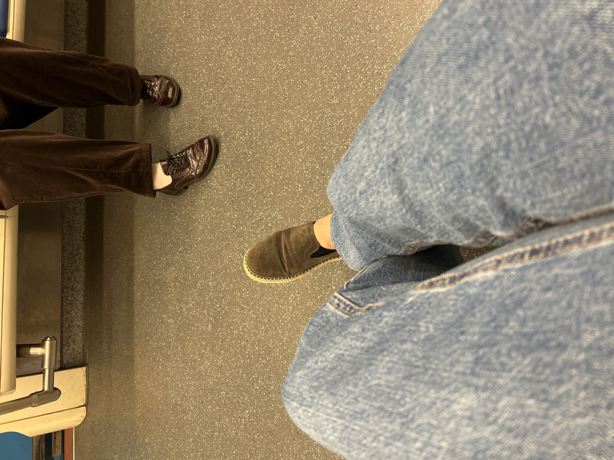The image shows a close-up of two people's legs and feet on the floor of a public transportation vehicle, likely a train or bus. The floor is a speckled gray material. One person, in the foreground, is wearing light blue denim jeans and a brown suede slip-on shoe. The other person, in the background, is wearing dark brown corduroy pants and brown leather dress shoes. The feet of the person in the background are closer together, suggesting they are sitting or standing in a more conventional posture, while the person in the foreground has their legs extended. The environment suggests an indoor setting with utilitarian flooring and seating structures that are partially visible at the top of the frame. There is no indication of the location within Moscow, Russia.