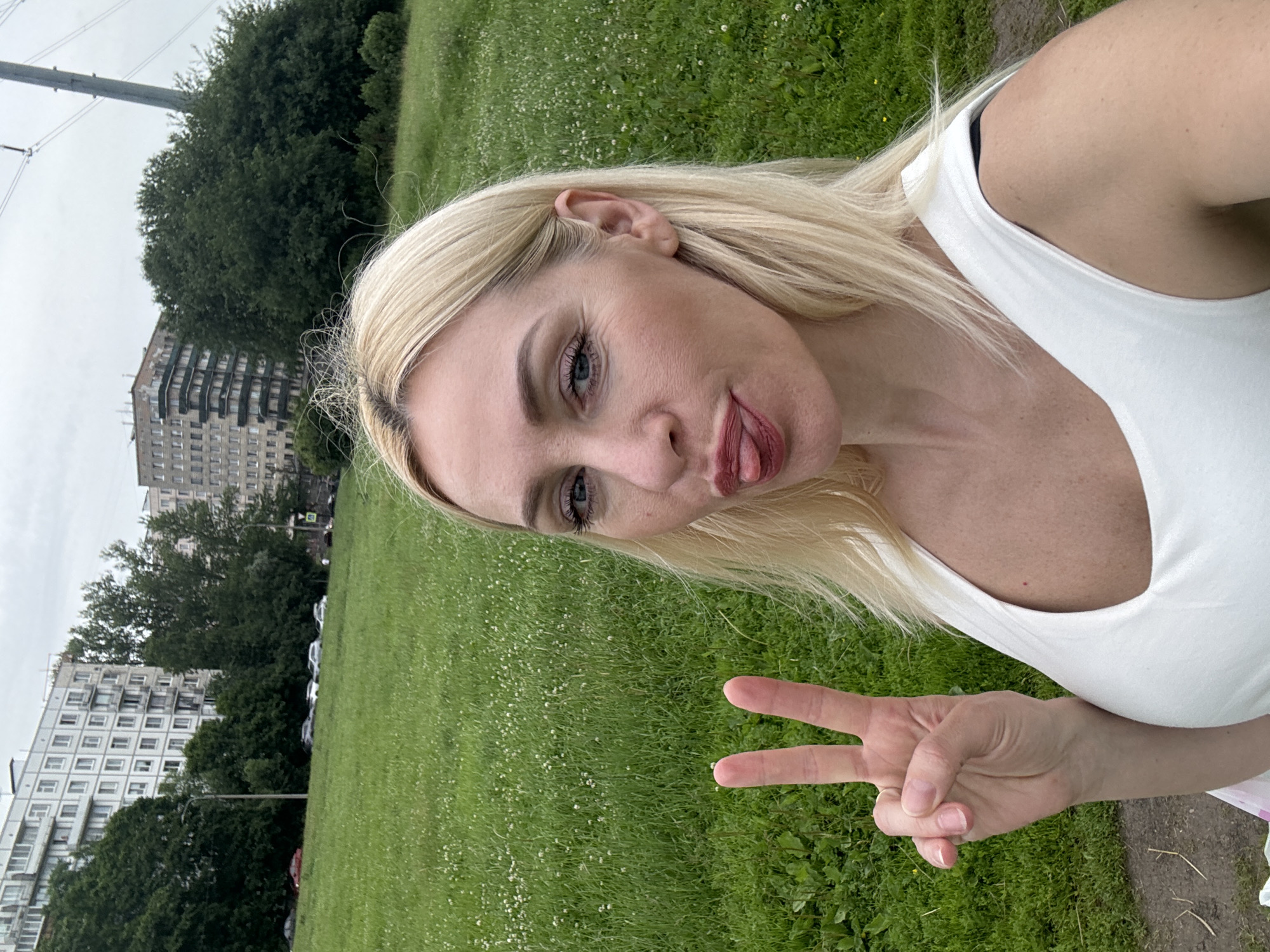 A blonde woman with fair skin, wearing a white tank top, stands in a grassy field with a slightly overcast sky. She is making a peace sign with her left hand and sticking her tongue out. Behind her, there are trees and tall, multi-story apartment buildings, some of which appear to be under construction. Cars are parked in the distance. The grass is green and dotted with small white flowers. A power line pole is visible in the background.