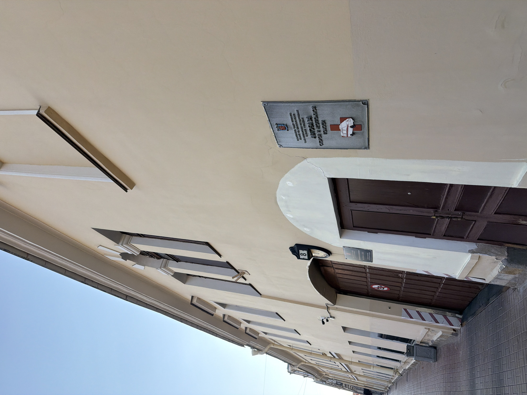 The image depicts the exterior of a building on a sunny day. The building is light yellow with a light-colored cornice and arched windows with dark frames. There are two large, dark brown doors visible. One door has a circular sign with a red "no entry" symbol, and the other door is closer to the viewer. Above the second door, a metal plaque is mounted on the wall. The plaque features text in Cyrillic and a symbol of an ambulance with a red cross. A number "8" is displayed on a black sign above the circular "no entry" sign. The ground in front of the building is paved with cobblestones. The sky is clear and blue. The building appears to be an older structure located in Saint Petersburg, Russia, likely housing an ambulance station, as indicated by the plaque.