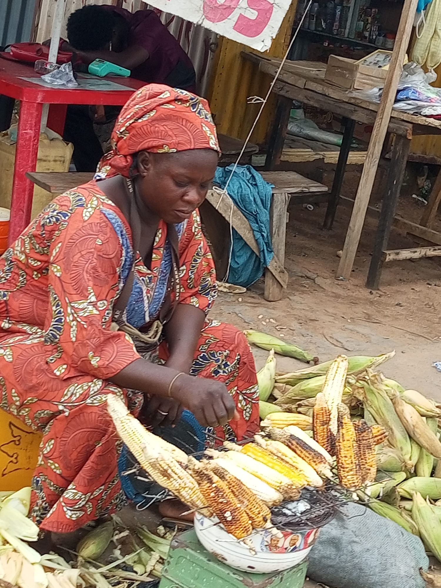 A full-shot, medium-angle outdoor photograph depicts a woman in the city of Tempure, Nigeria, seated on the ground and attending to roasted ears of corn. The woman, wearing a red headwrap and a red and blue patterned dress, is focused on the corn laid out in front of her on a metal grate, which is placed in a decorative bowl. The corn appears to be in various stages of roasting, with some kernels golden yellow and others charred black. Unroasted ears of corn, still in their husks, are piled around her. The ground is sandy and dusty, indicative of a dry outdoor environment. In the background, a stall is visible with shelves stocked with items, and wooden tables are set up, suggesting a market setting. Another person, partially visible, is seated at a red table in the background. The overall scene conveys a busy, open-air market atmosphere.