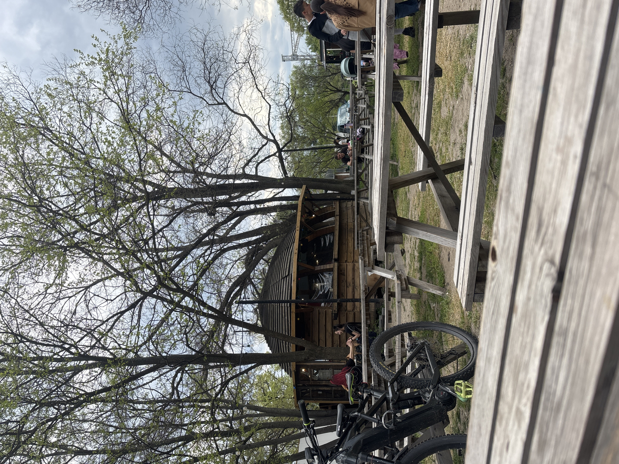 This image depicts an outdoor recreational area, likely in Plovdiv, Bulgaria, under an overcast sky during daytime. The scene is characterized by numerous wooden picnic tables, a rustic wooden cabin-like structure, and several large trees, some showing the first signs of spring with new green leaves while others remain bare.
In the foreground, a black bicycle with distinctive bright yellow pedals is prominently featured, positioned near a weathered wooden picnic table that extends from the right into the mid-ground.
The mid-ground centers around a two-story wooden building, resembling a cabin or kiosk, nestled among the tree trunks. Several people are visible throughout the area; two individuals are seated at a picnic table close to the wooden structure, one with red hair wearing a red top and another in a black top, appearing to be conversing. Further back, more people are seated at other picnic tables, contributing to a casual social atmosphere. Towards the right side, a group of people stands near more tables, with a baby car seat or stroller visible on one of them.
In the background, a white vehicle is partially seen, and a large metal scaffolding structure, possibly indicating an outdoor stage or event setup, rises above the tree line. The ground consists of dirt and sparse grass. The overall impression is one of a relaxed gathering spot, possibly an outdoor eatery or event space. No discernible text is visible in the image.