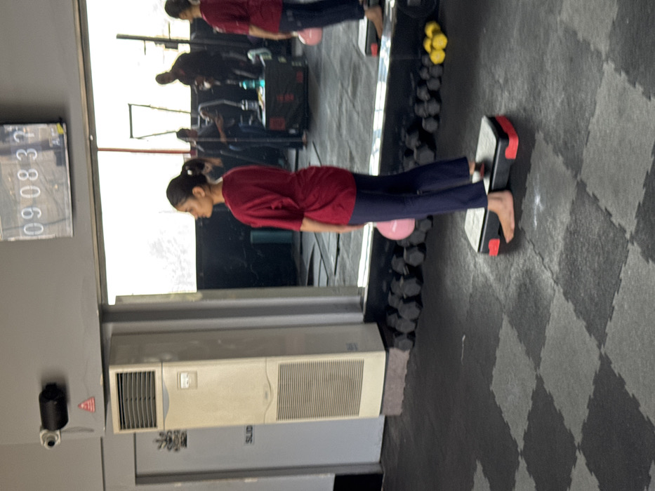 A person is captured in a gym setting, standing with their feet elevated on a step platform and a pink ball positioned beneath their lower abdomen. Their posture suggests a balance exercise. The reflection in a mirror shows other people in the background. Gym equipment like dumbbells are visible near the mirror and the floor has a black and gray patterned mat. A security camera and a large white air conditioning unit are mounted on the wall.
