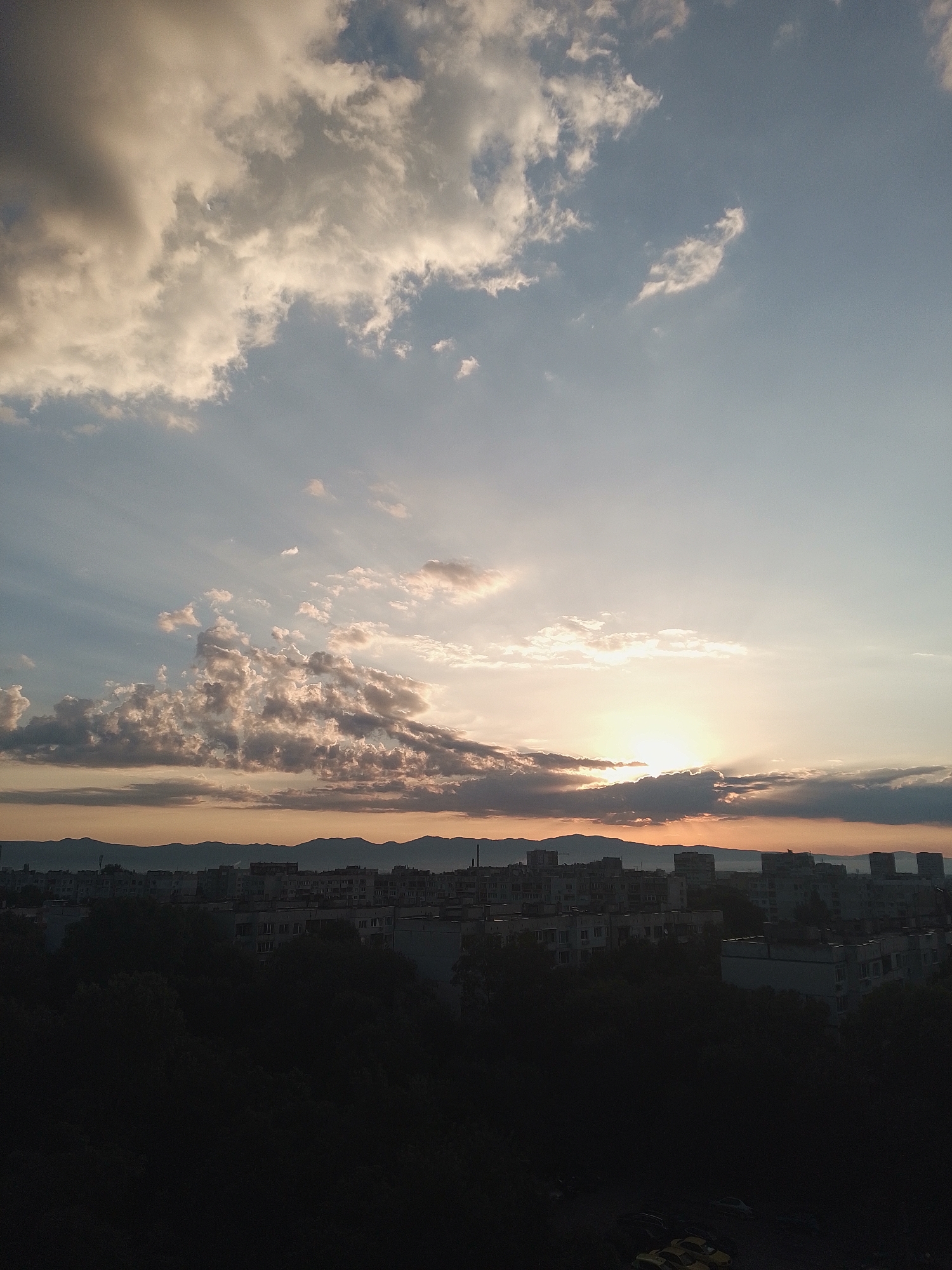 The image shows a cityscape at sunset, possibly from an elevated viewpoint in Sofia, Bulgaria. The sky is filled with dramatic clouds, with the sun partially obscured by them, casting a warm glow over the scene. In the foreground, a silhouette of trees and buildings is visible. The middle ground features a dense collection of residential buildings, characteristic of urban areas in Sofia. The background shows a mountain range, likely the Vitosha Mountain, which borders Sofia to the south. The overall impression is a serene urban sunset with natural and man-made elements harmoniously blended.