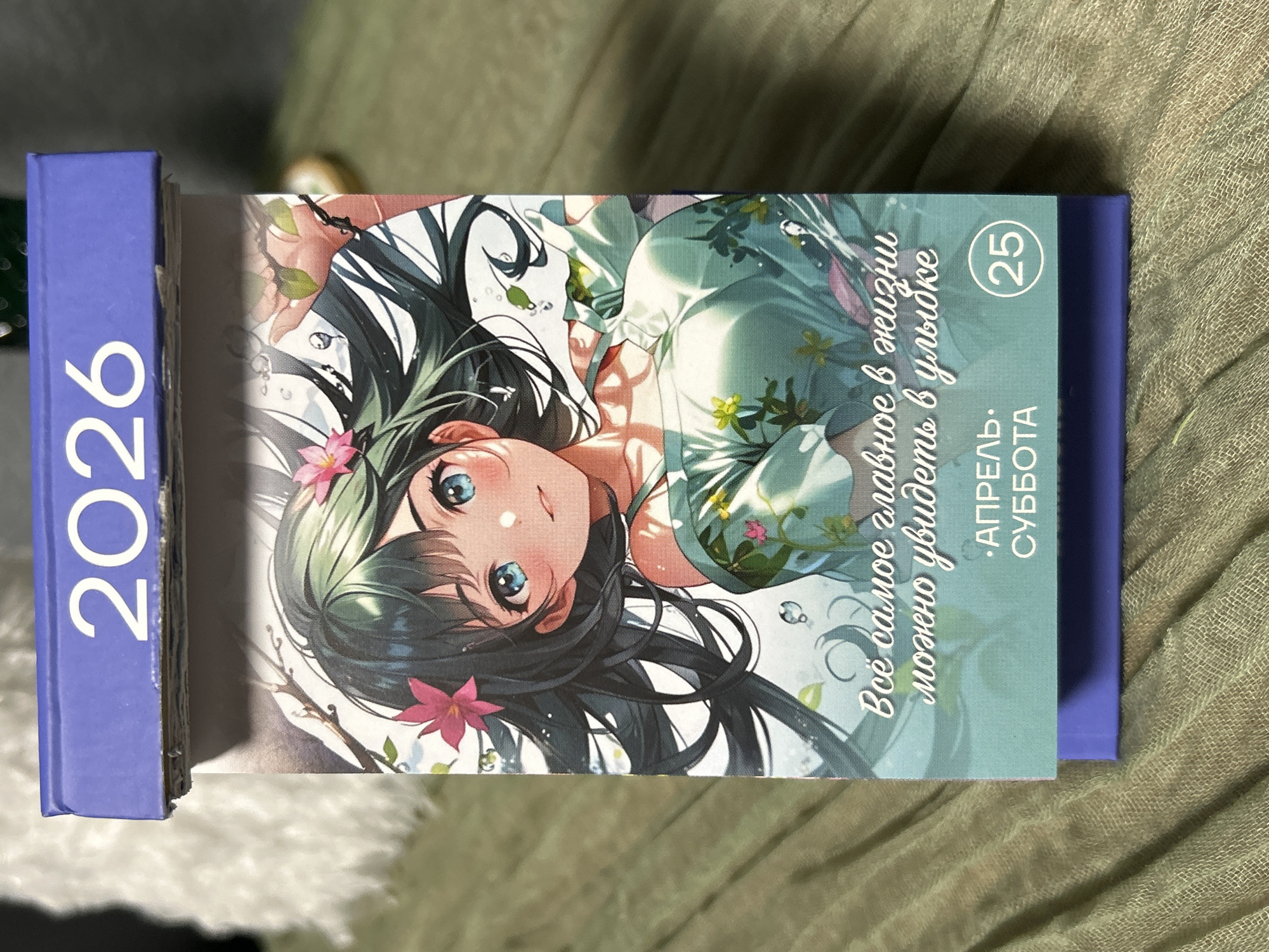 A 2026 desk calendar is shown. The calendar is blue, with the year "2026" printed vertically in white. The calendar is lying next to a book with a blue cover. The cover features an anime-style illustration of a girl with black hair, blue eyes, and pink flowers in her hair. The text on the cover is in Russian, "Всё самое главное в жизни можно увидеть в улыбке" which translates to "The most important thing in life can be seen in a smile." The date "25" is circled and the text below translates to "April, Saturday". The background has a textured green surface.