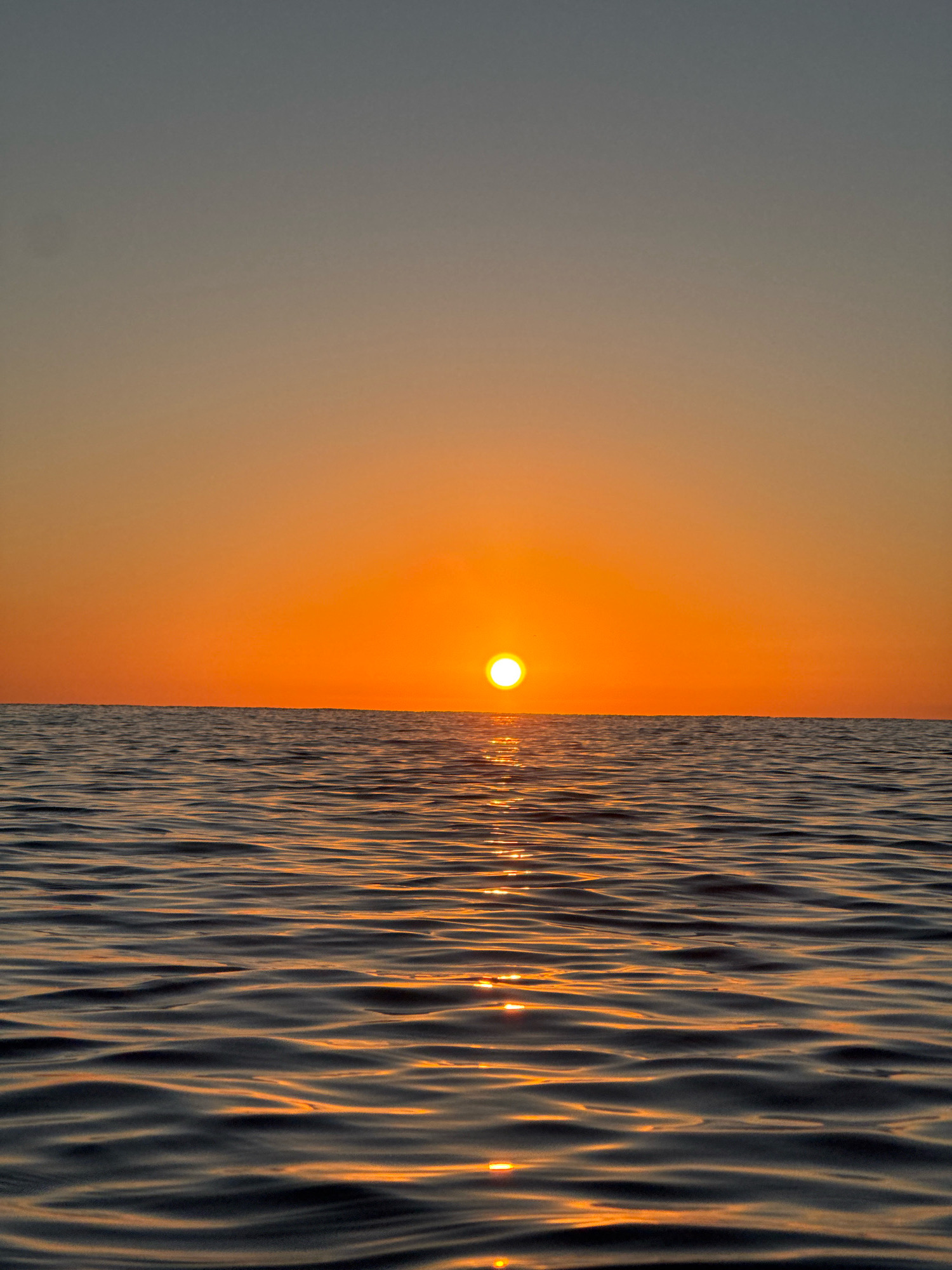 The image depicts a serene seascape at sunset, with the sun a bright orange orb resting just above the horizon. The sky transitions from a soft grey at the top to a warm, vibrant orange as it approaches the sea. The water's surface is rippled, reflecting the colors of the sky and the sun's brilliant glow in scattered streaks of light. The horizon line is sharp and clear, separating the sky from the dark, wavy expanse of the ocean. The scene evokes a sense of tranquility. The location is the Mediterranean Sea off the coast of Calp, Spain, with the iconic Ifach Rock likely forming part of the skyline or seascape, though not explicitly visible in this particular frame.