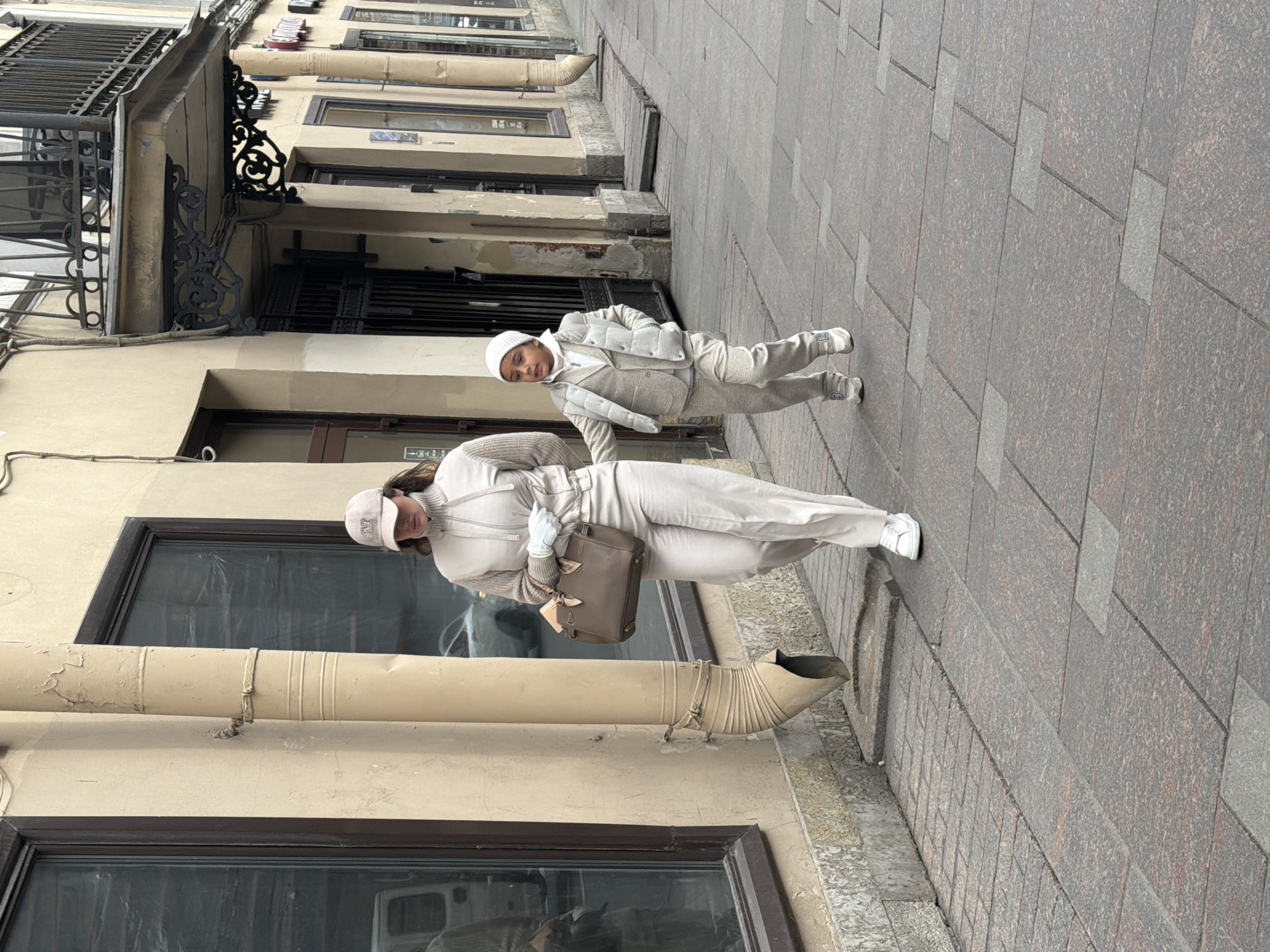 A street scene shows two people on a sidewalk alongside an old building. The building's architecture is characterized by large windows, dark window frames, and decorative ironwork on the upper floors. A beige drainage pipe is attached to the building.
The sidewalk is paved with large, rectangular stone slabs. One person, an adult wearing a white outfit, a white hat, and carrying a brown satchel, walks along the sidewalk. A younger child, also wearing a white outfit and hat, walks slightly ahead of the adult. They are on a street in Saint Petersburg, Russia.