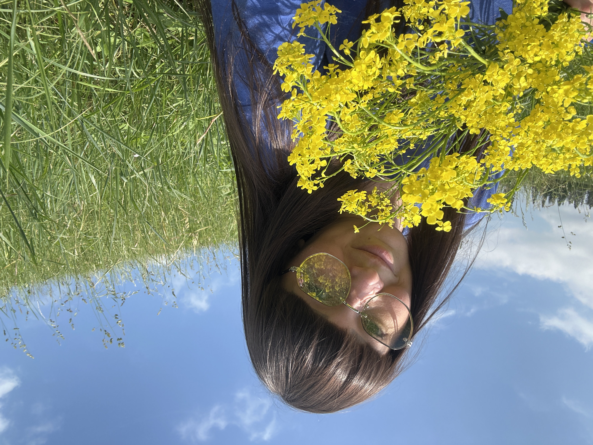A young woman with dark hair and bangs, wearing a blue shirt and sunglasses, holds a bouquet of bright yellow flowers. She is outdoors, in a grassy field under a clear blue sky with scattered white clouds. The sun is shining, casting reflections of the yellow flowers on her sunglasses. The grass is tall and green, and some trees can be seen in the distance. The overall impression is one of a pleasant summer day. The location is specified as Buranovo, Russia.