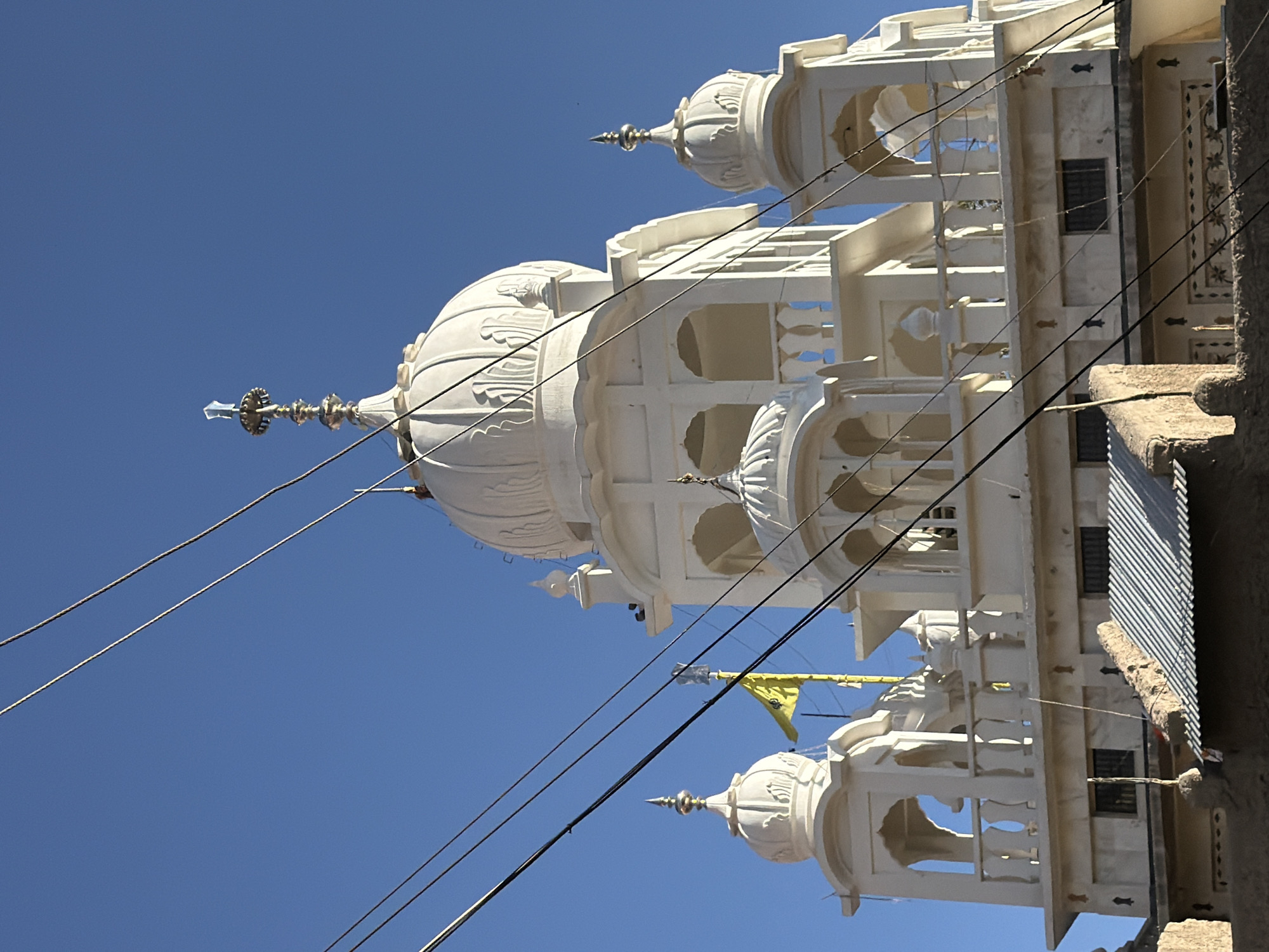 A white building with domes and intricate architectural details is shown against a clear blue sky. The building appears to be a temple or a similar religious structure, possibly a Gurdwara, given the presence of a yellow flag with a religious symbol, which is likely the Khanda. The structure has multiple levels, arched windows, and decorative finials on its domes and spires. Power lines are strung across the foreground, partially obscuring the building. Below the main structure, there are elements of simpler, more rustic construction with corrugated metal roofs and mud or stone walls. The context of Leh, India, suggests this might be a location within or near the city, a region known for its Buddhist monasteries and Tibetan-influenced architecture, although this particular structure appears to have Sikh or other influences. No people are visible in the image. The lighting suggests it is daytime, possibly morning or afternoon, with the sun casting shadows.