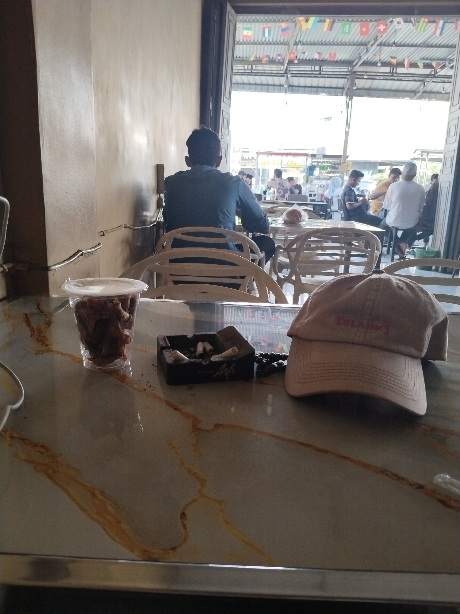 The image captures a scene at a food stall or eatery in Rawang Itiek, Indonesia, focusing on a table in the foreground and a broader view of the establishment through an open doorway. On the table, a clear plastic cup filled with a dark-colored, possibly food-related substance, sits next to a black ashtray containing several cigarette butts. Adjacent to these items, a light brown baseball cap with the embroidered text "[BLANK]" lies on its side. The table itself has a marbled surface with prominent brown streaks. In the background, through the open entrance, a bustling outdoor seating area is visible with several tables and chairs occupied by people. Some patrons are seated, others appear to be standing or walking. The roof of the establishment is supported by metal beams and decorated with numerous small flags of various countries. The lighting suggests it is daytime, with sunlight filtering into the space. The overall impression is of a casual, public gathering place.