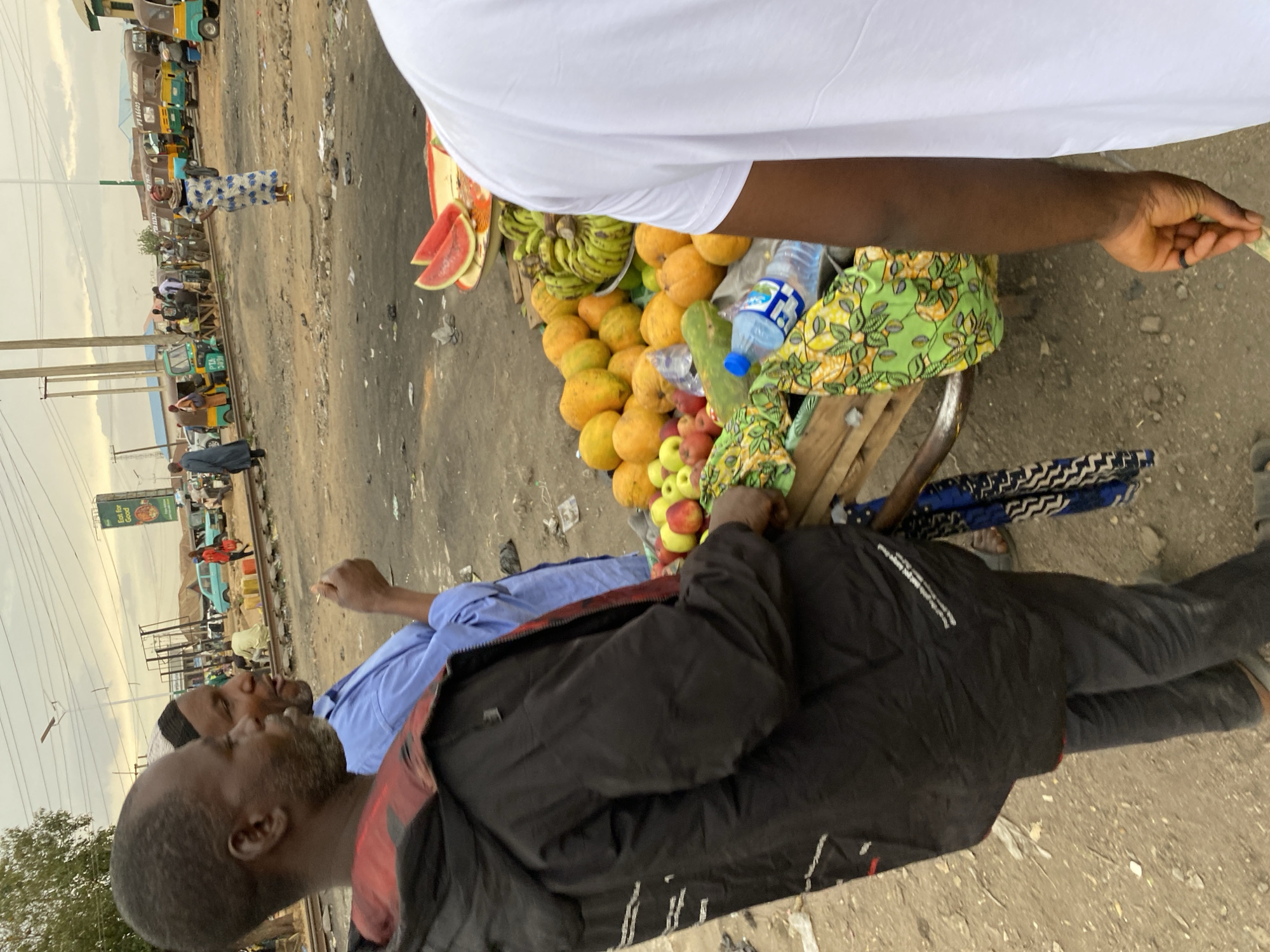 The image shows a street scene in Bukuru, Nigeria. A fruit vendor operates a cart laden with mangoes, bananas, watermelon, apples, and bottled water. A customer in a white shirt is making a transaction, handing the vendor currency. Another man in a black jacket and a light blue shirt is standing nearby talking to someone not entirely visible. Other people are walking or standing in the background along a railway track with a street running parallel to it. Various vehicles, including auto-rickshaws and cars, are present along the street. The ground is a mix of dirt, asphalt, and debris.