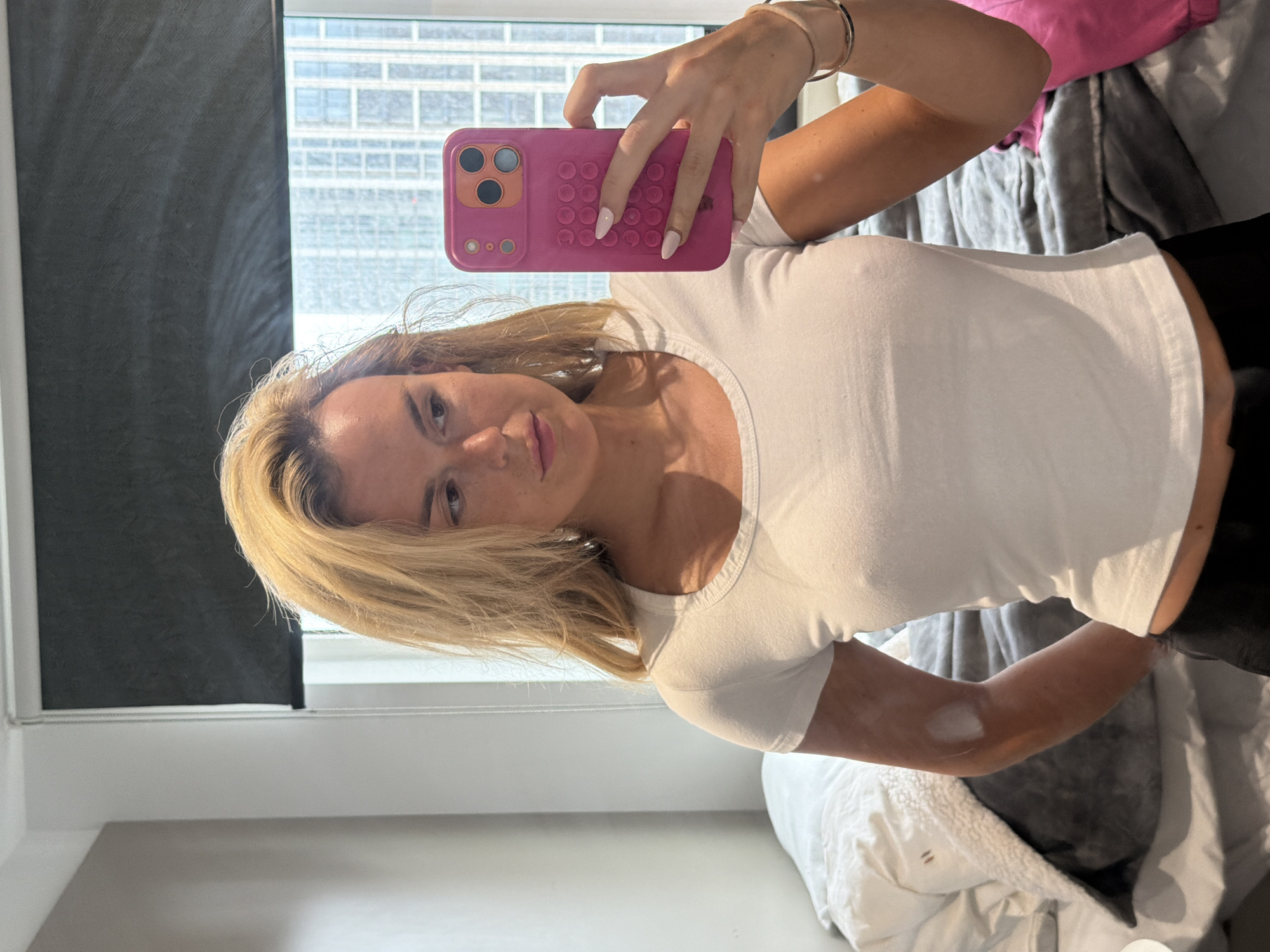 A young Caucasian woman with blonde hair is taking a selfie in a bedroom. She is wearing a white t-shirt and black pants. She is holding a pink phone in her right hand. The room appears to be an apartment in a high-rise building in London, as evidenced by the cityscape visible through the window behind her. The blinds are partially drawn, and there is a bed with grey and white bedding to her left. The woman has light skin with some uneven pigmentation on her left arm.