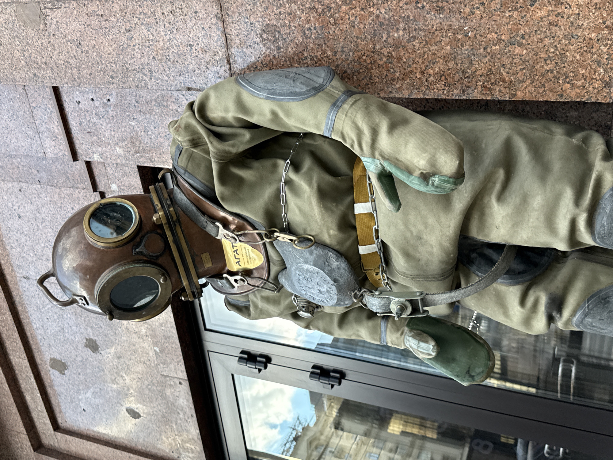A vintage deep-sea diving suit is displayed leaning against a building facade. The suit is a muted green color and appears to be made of heavy canvas or rubber. It features a large, round helmet with two glass portholes, a metallic breastplate with chains, and thick gloves. A yellow and white striped belt is cinched around the waist. A metal tag with Cyrillic lettering, possibly indicating a brand or model, is attached to the front of the suit. The building behind the suit is clad in reddish-brown granite and has a large glass storefront to the left, reflecting a cloudy sky and buildings. The overall scene suggests a display, possibly outside a museum or historical exhibit, in a city environment. The text on the tag reads "АГАТ" and "ЗАБАСТОЕНОСКЕИ НАСОСНОИ МАРКЕ". The suit is positioned on a sidewalk.