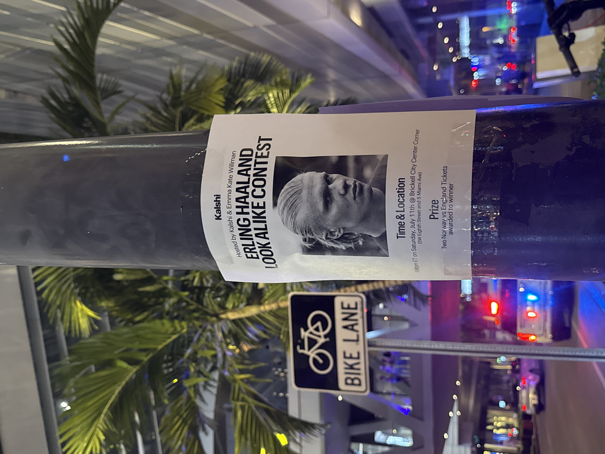A black lamp post in an urban environment displays a flyer for an "Erling Haaland Look Alike Contest." The flyer features a black and white portrait of a man with blond hair, presumably Erling Haaland, and text detailing the event's hosts, time, location, and prize. Below the flyer, a "BIKE LANE" sign is visible. The background shows a street scene at night, with palm trees, blurred figures, a police car with flashing lights, and illuminated buildings. The context of Brickell City Center and Miami Avenue indicates the location is Brickell, Miami, Florida, USA.